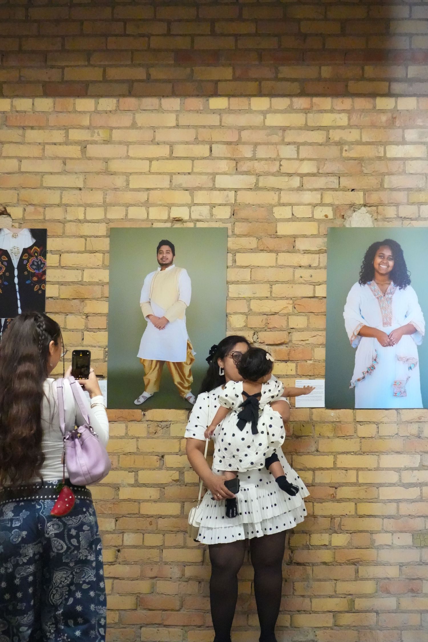 Two women pose in front of portrait photos displayed on a brick wall at an art exhibition.