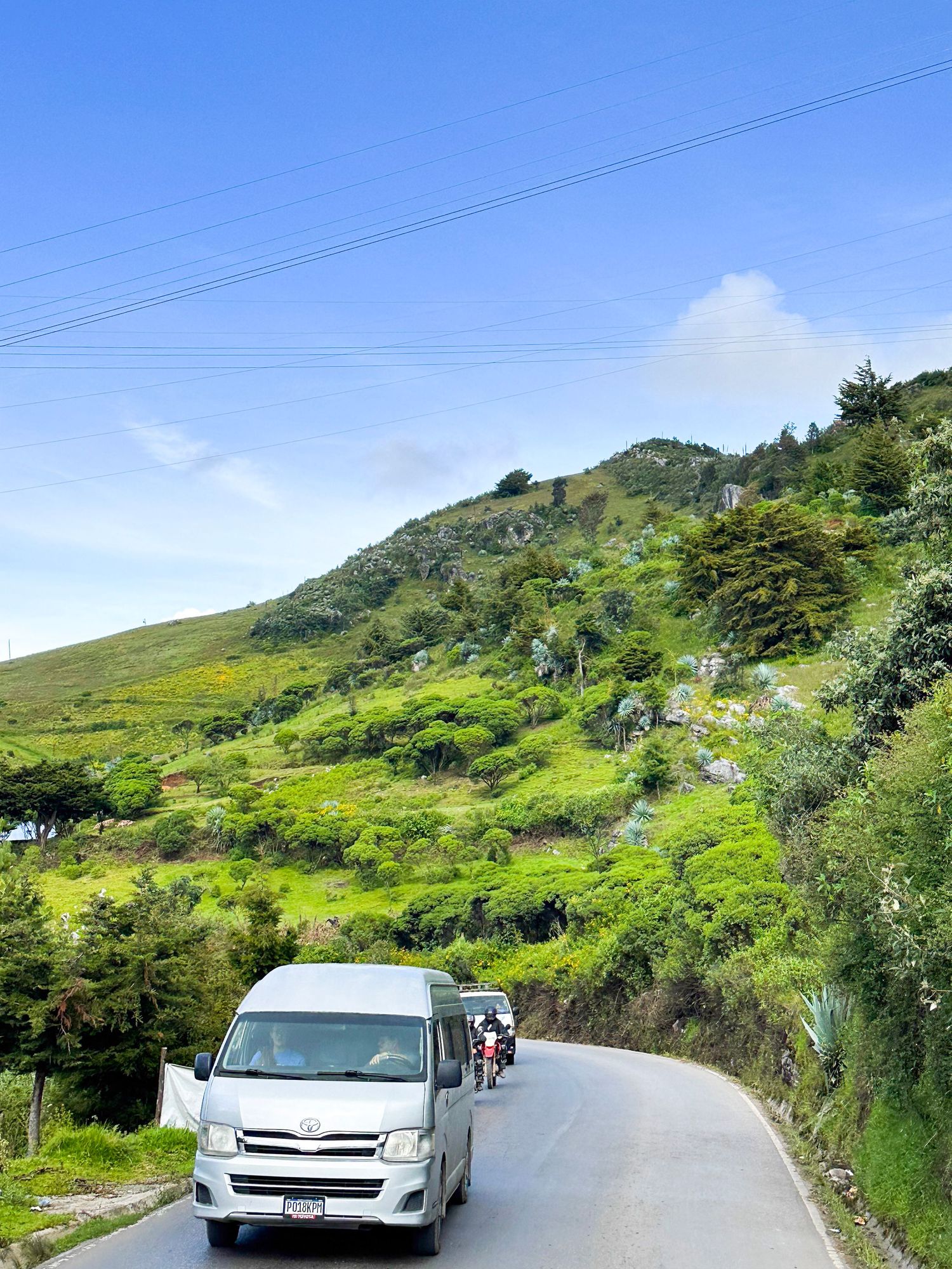 White vehicles drive along a winding mountain road with lush green hillsides and blue sky in the background, Guatemala, travel photography