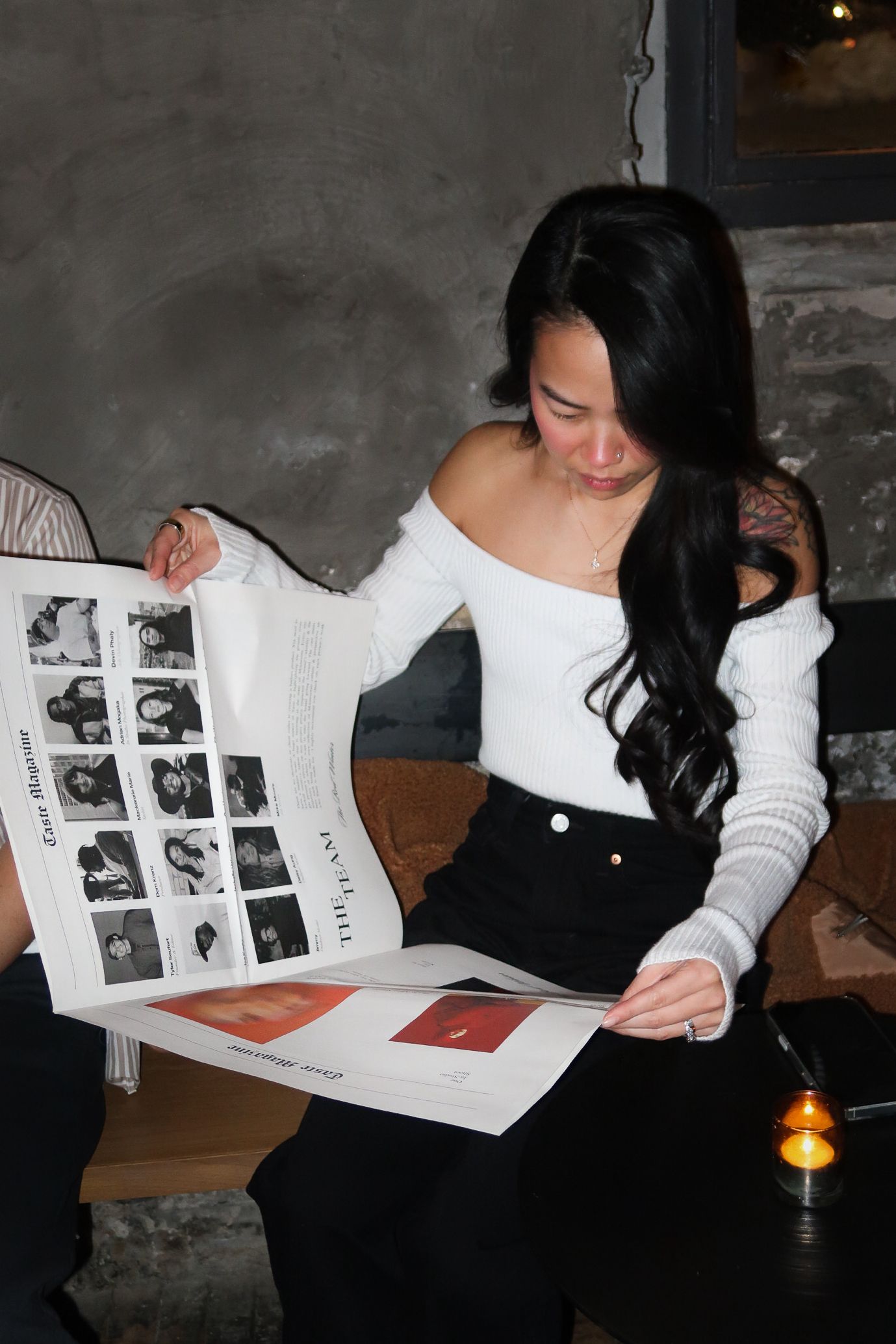 Young woman with long dark hair in white off-shoulder top reading a magazine while seated indoors.