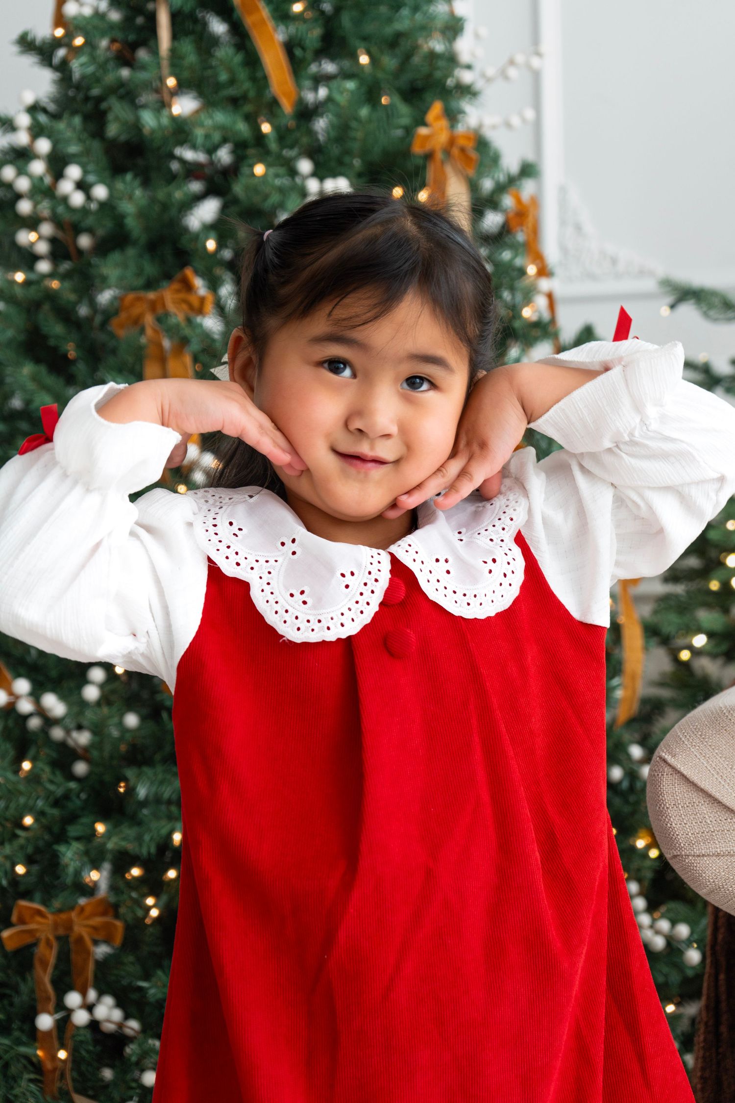Young Asian girl in red velvet dress poses in front of decorated Christmas tree with golden ribbons.
