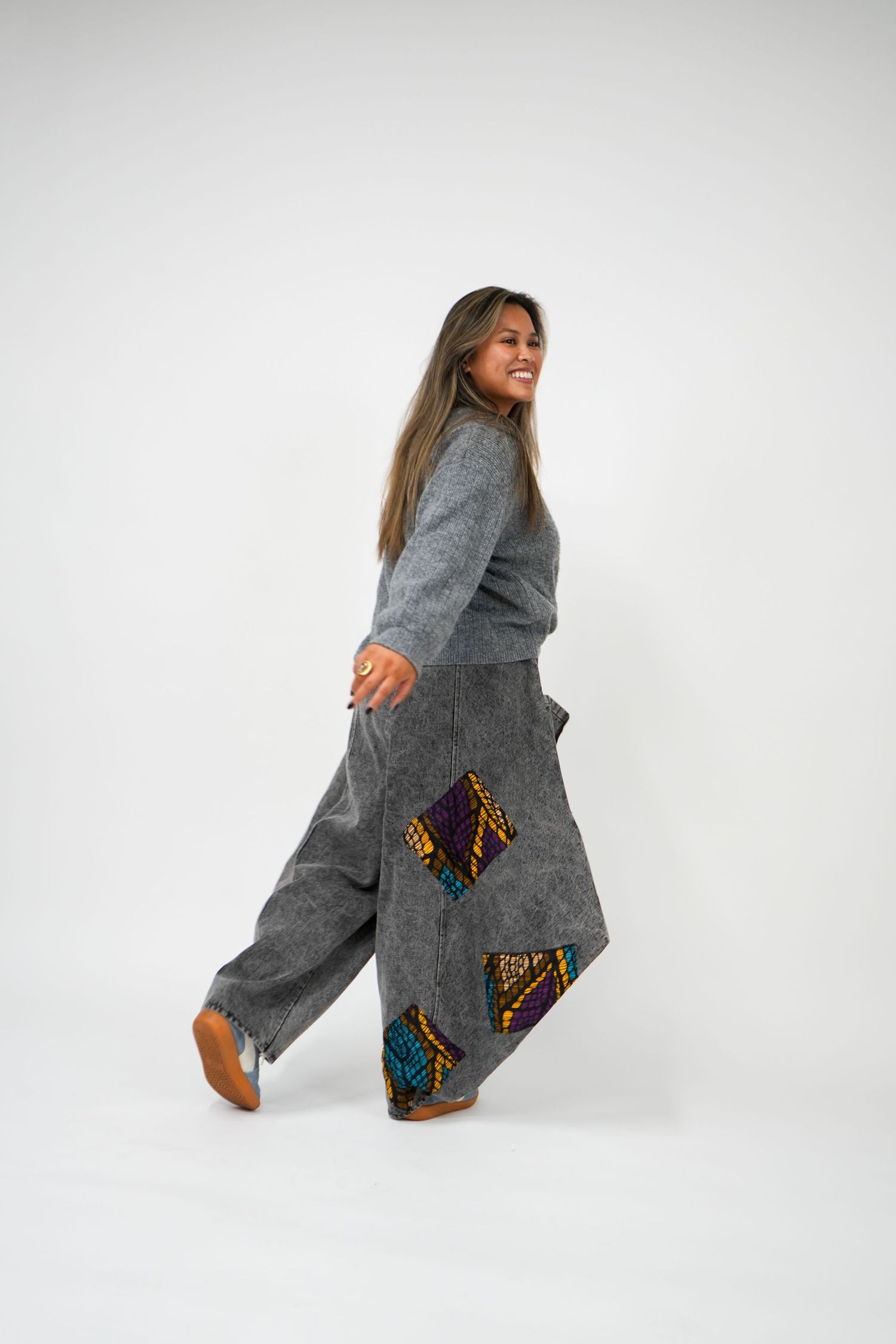 Woman in gray sweater and wide-leg patchwork pants with colorful geometric patches, walking and smiling.