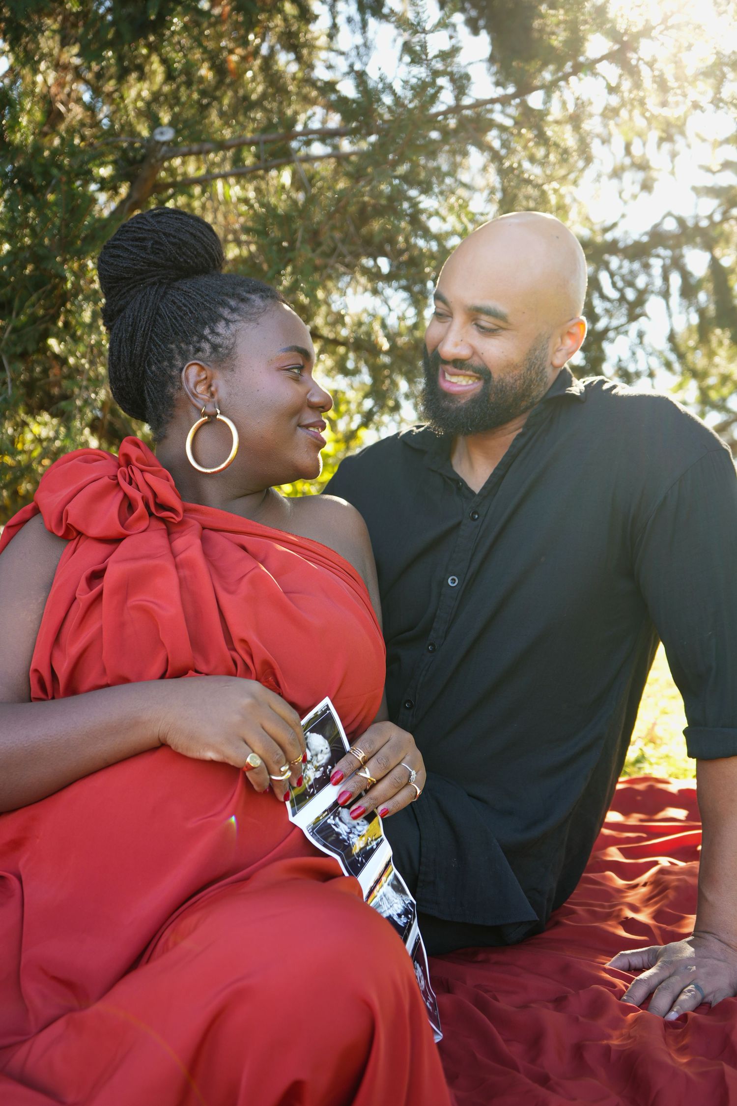Happy couple in red outfits sitting outdoors, woman holding ultrasound photos, celebrating pregnancy together.