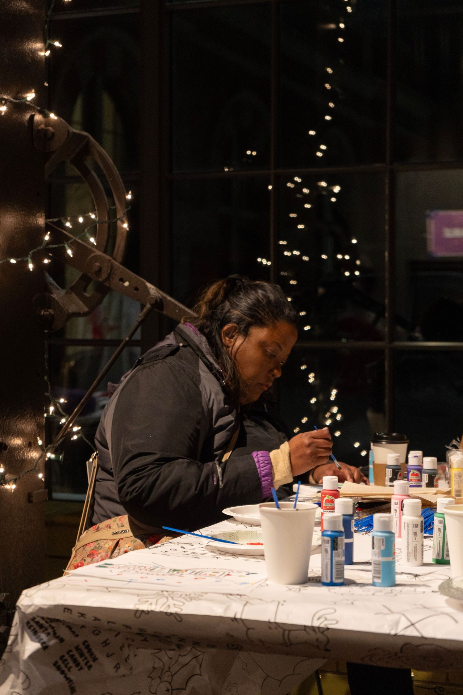 Woman painting at a table with craft supplies, surrounded by twinkling fairy lights in a dark indoor setting.