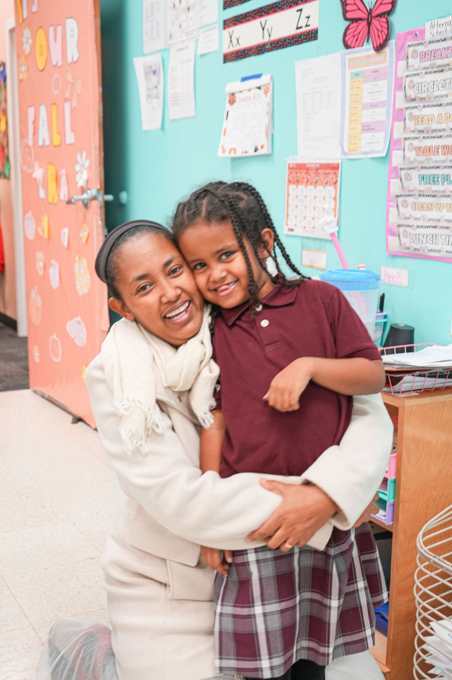 A woman and young girl in school uniform share a warm hug in a colorful classroom setting.
