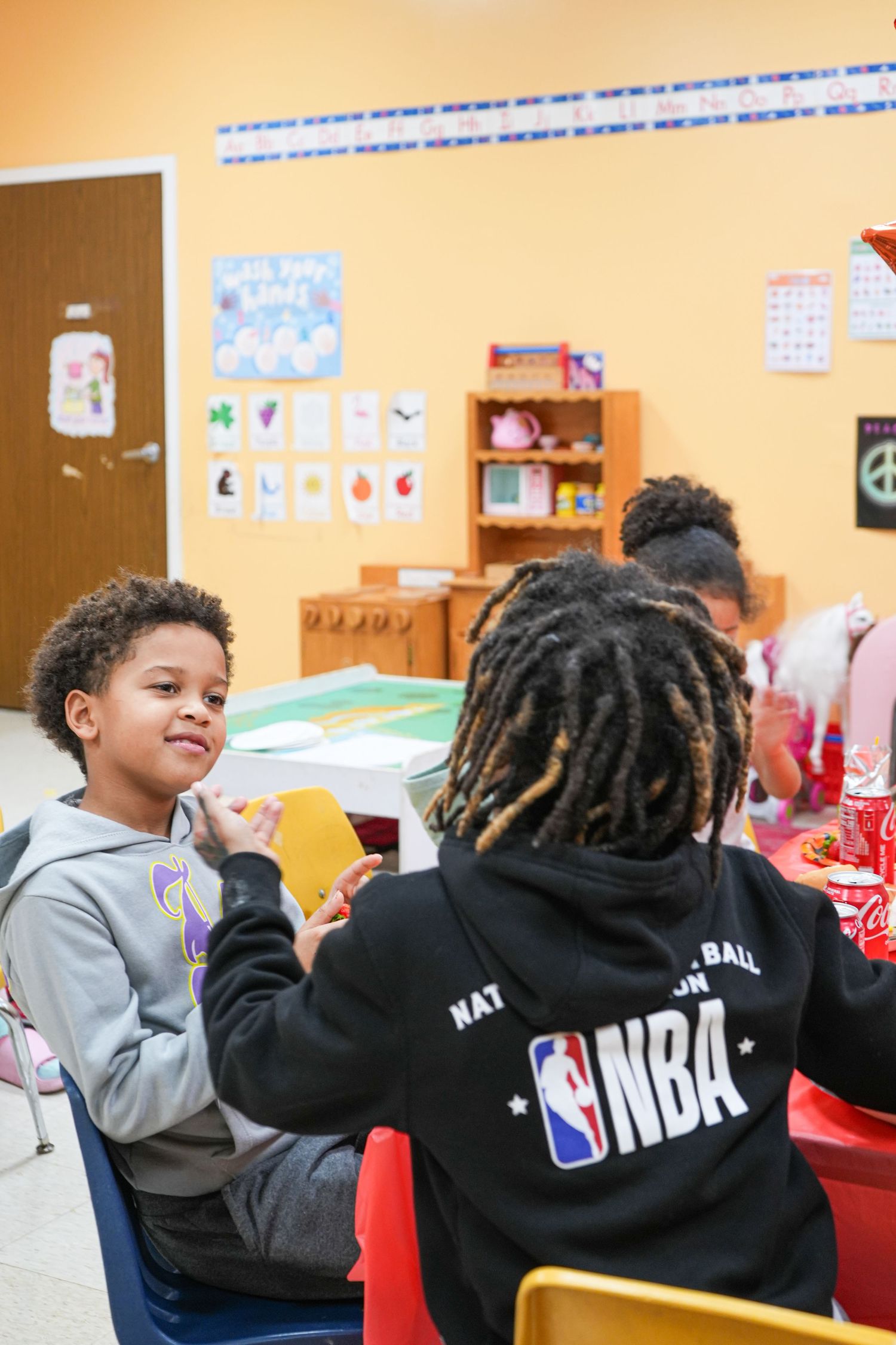 Two children sit together in a colorful classroom, one wearing an NBA hoodie, engaged in a fun activity.
