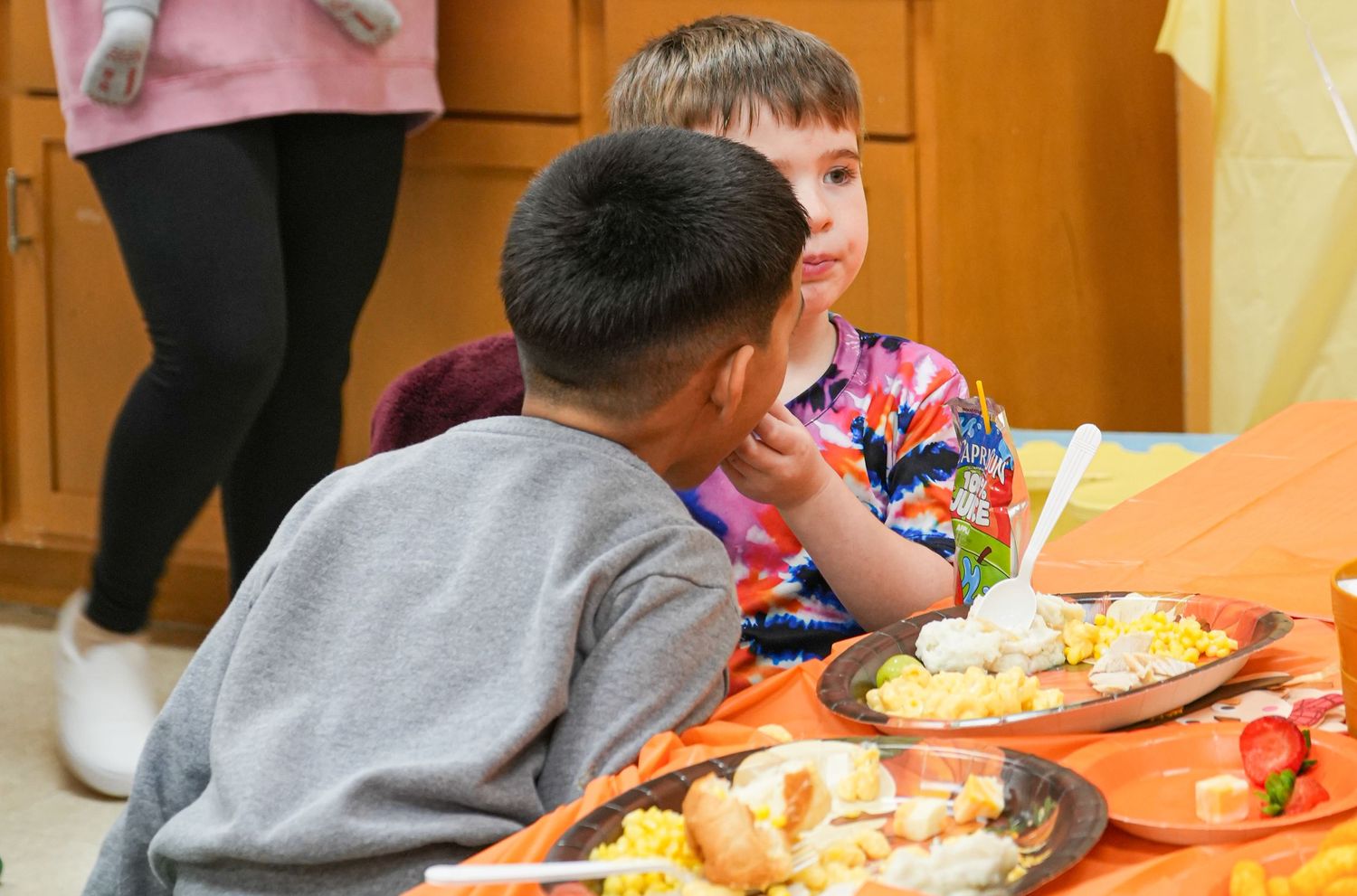 Two young boys sit together at a festive table with colorful dishes of food during a school celebration.