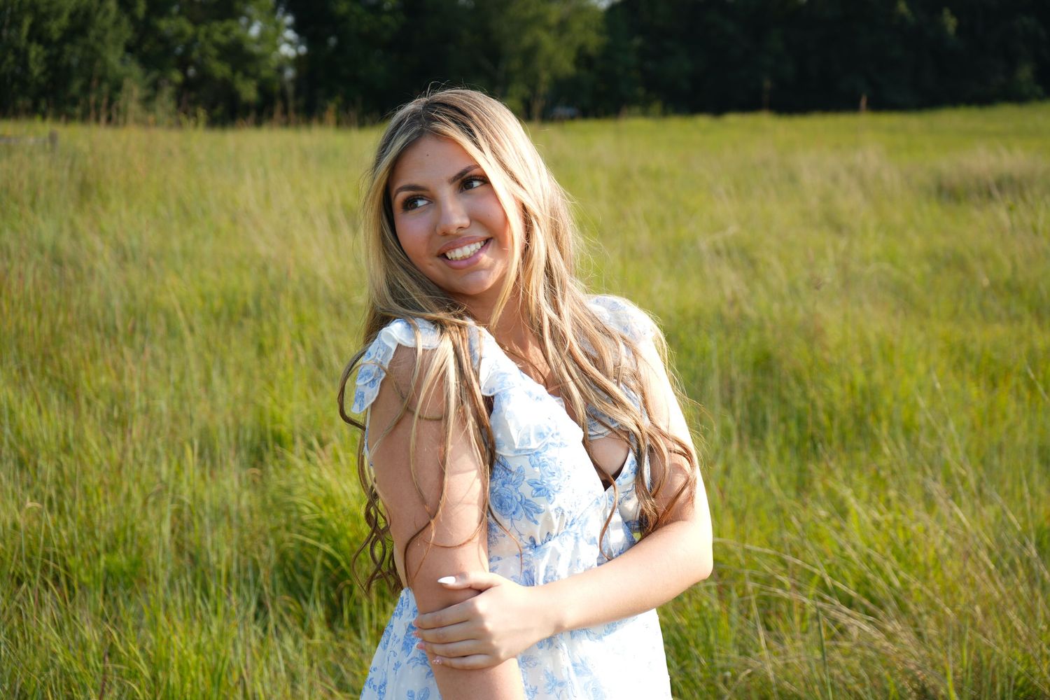 Smiling blonde woman in white floral dress holding boots in a sunny green meadow.