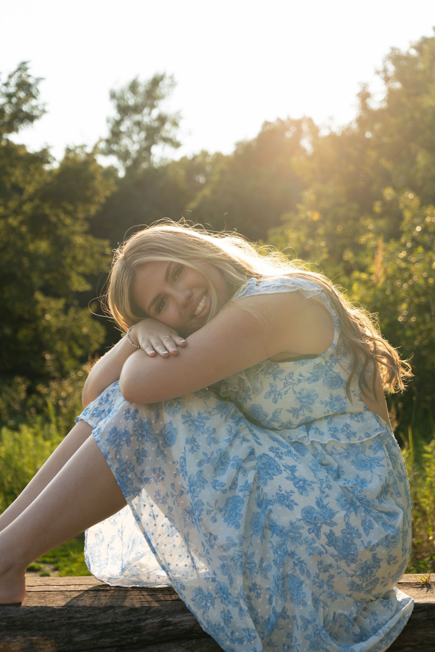Smiling blonde woman in blue floral dress sitting outdoors on log with golden sunlight and green trees behind her.
