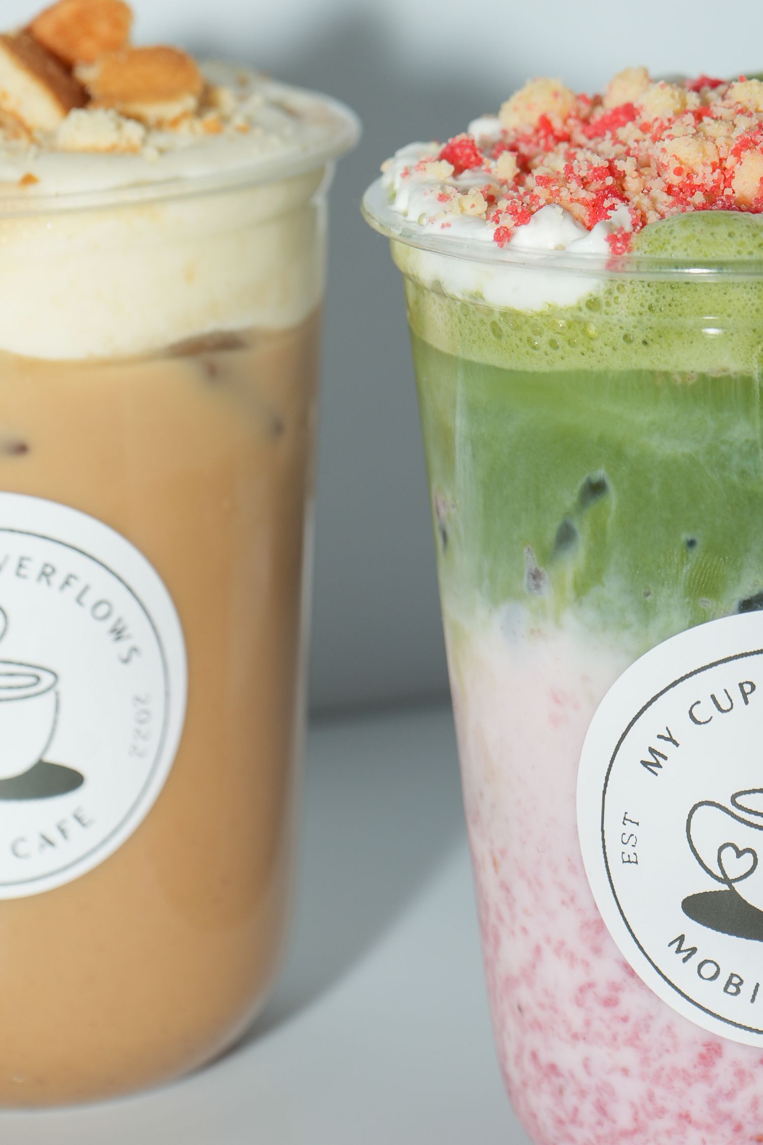 Two layered iced drinks from My Cup Overflows Cafe, featuring a matcha strawberry and a caramel coffee topped with crumbles.