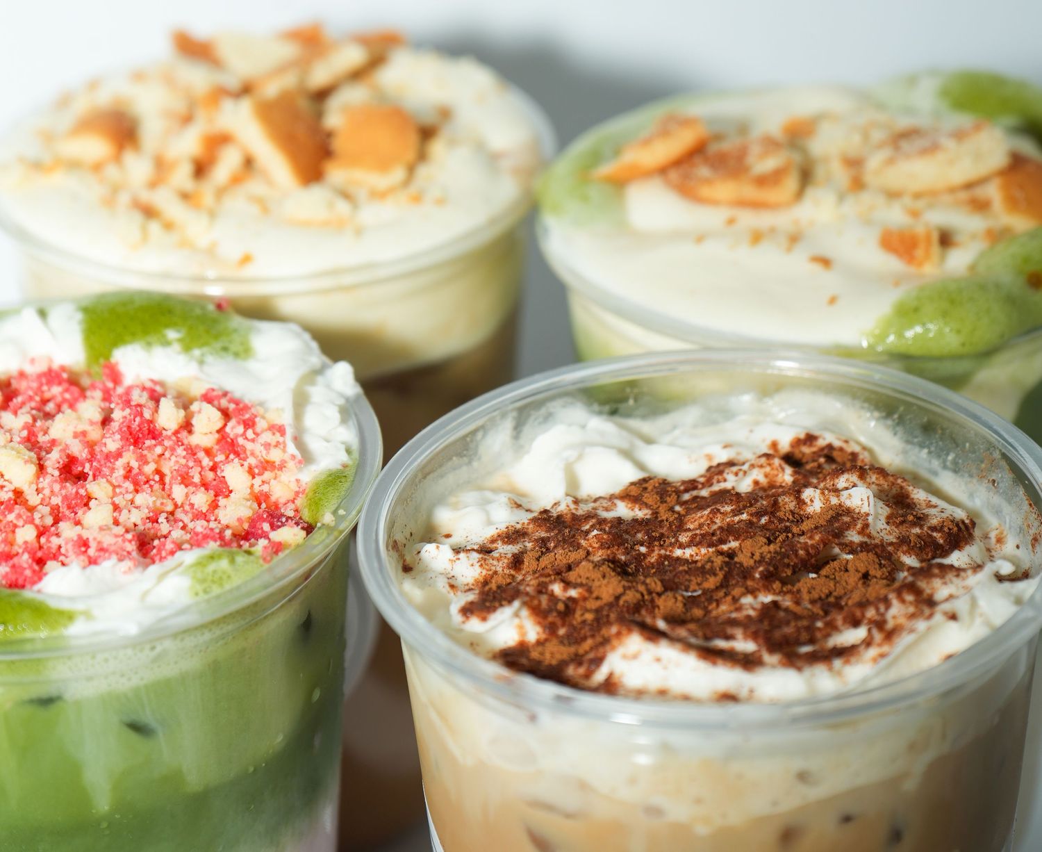 Four plastic cups filled with creamy desserts topped with whipped cream, chocolate shavings, crushed cookies, and fresh fruit.