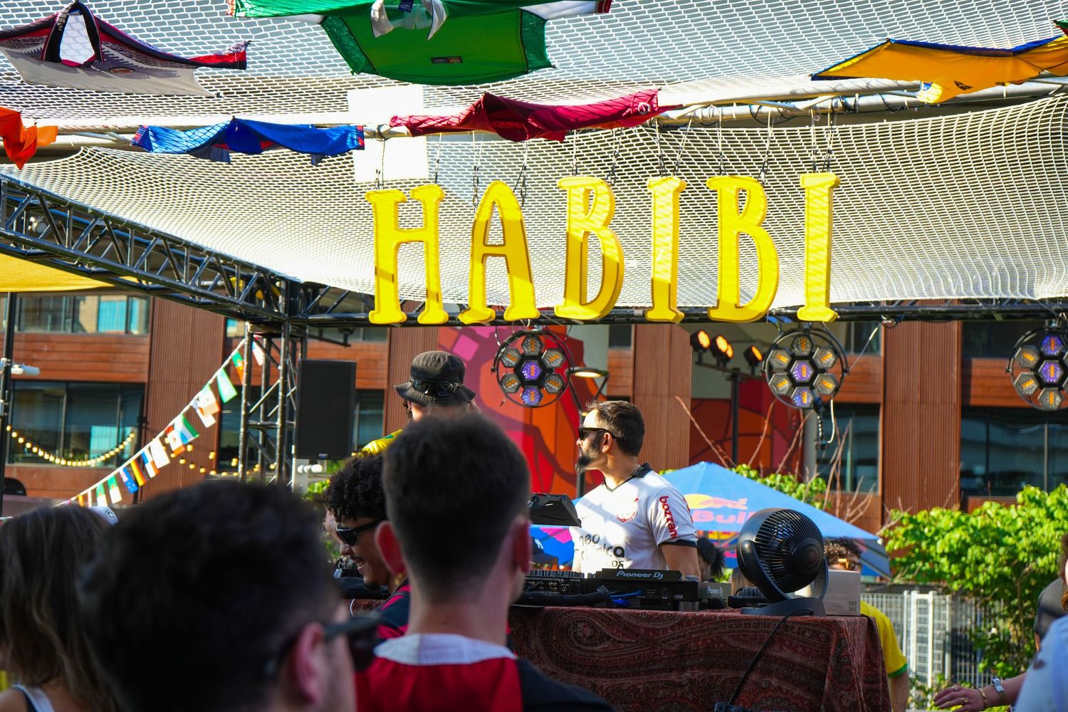 Colorful outdoor event with yellow HABIBI balloon letters sign above a DJ booth under decorative canopy.