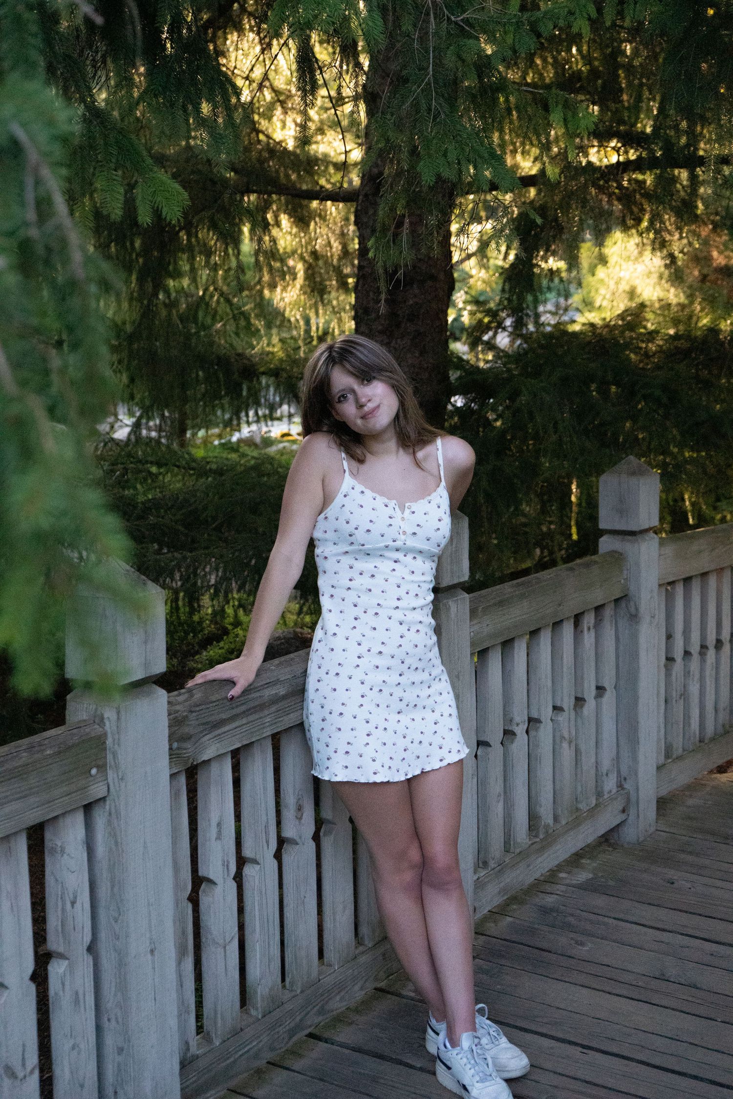 Young woman in white floral sundress posing on wooden bridge surrounded by lush green pine trees.