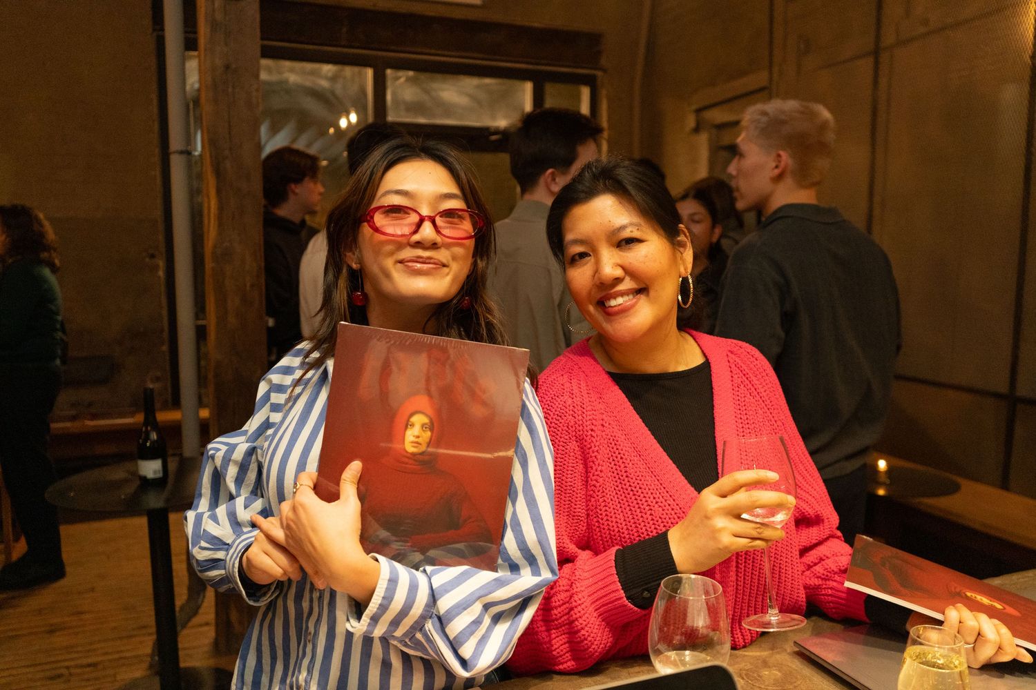 Two women smiling at a social event, one holding a red book, wearing colorful outfits in a warmly lit venue.