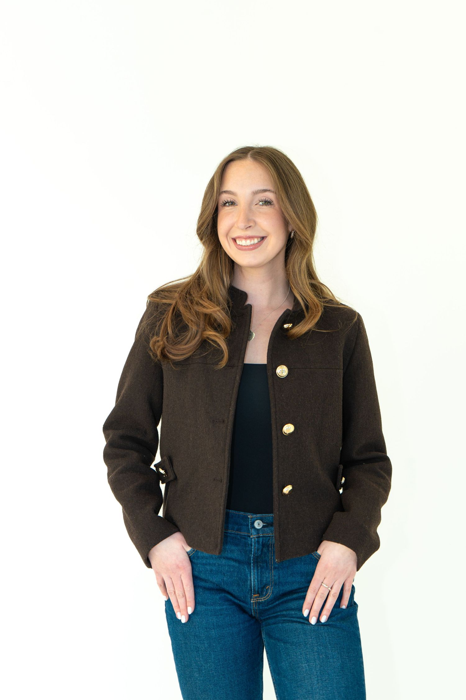 Woman smiling in brown button-up jacket, black top, and blue jeans against white background.