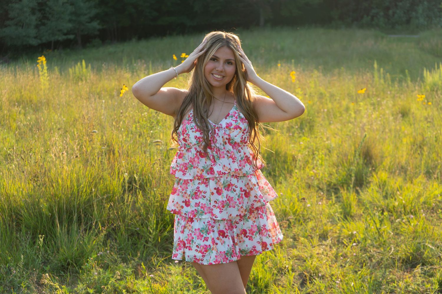 Young woman in floral dress posing in sunny meadow with hands in hair, smiling outdoors.