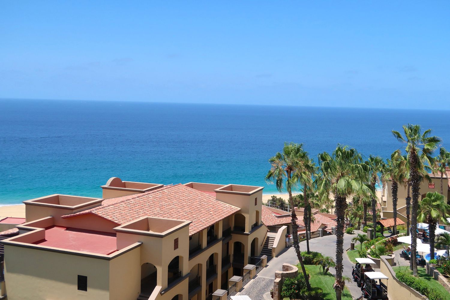Aerial view of a luxury beachfront resort with red-roofed buildings, palm trees, and turquoise ocean in Los Cabos, Mexico.