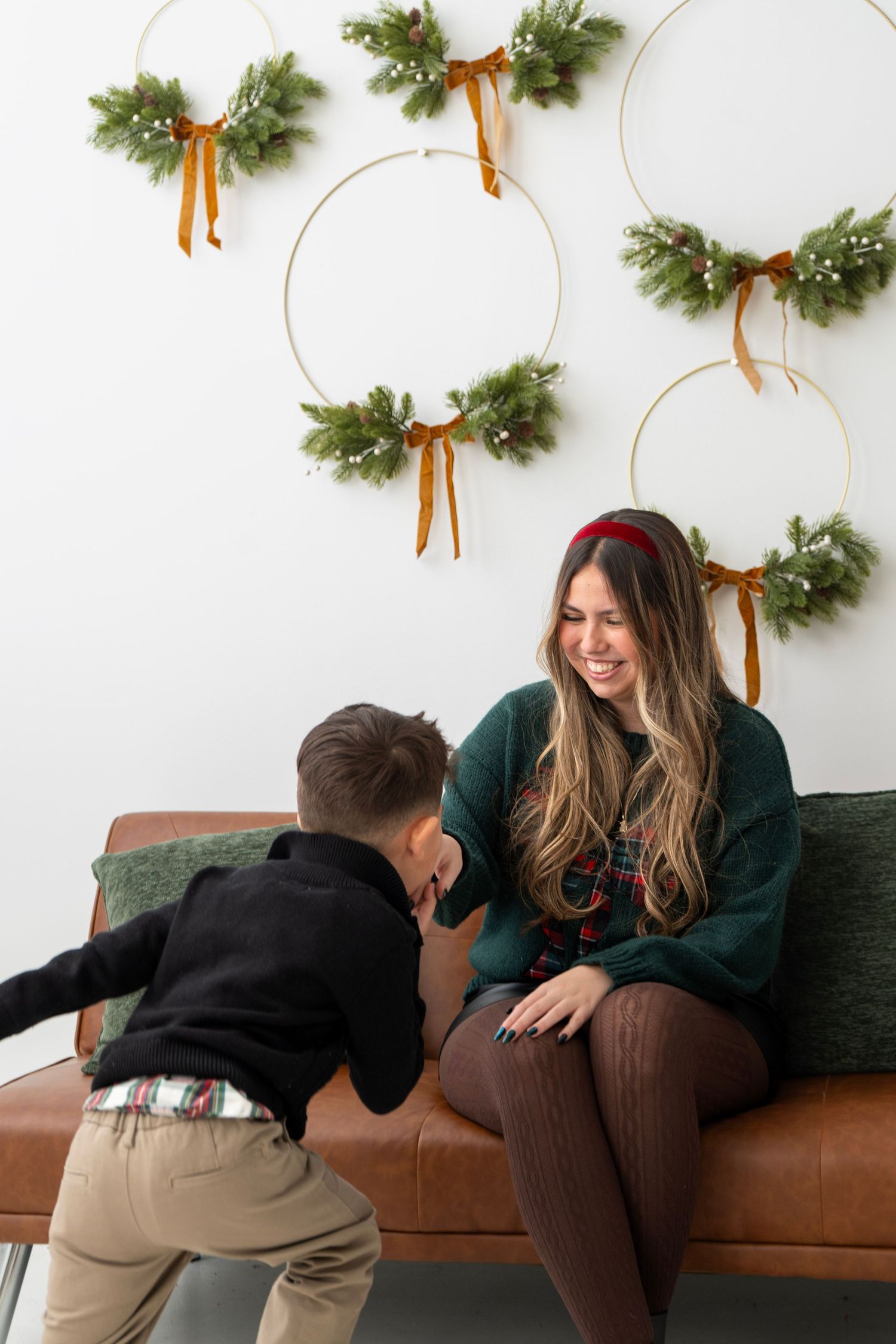A woman in a green sweater sits on a leather couch laughing with a young boy, surrounded by Christmas wreaths.