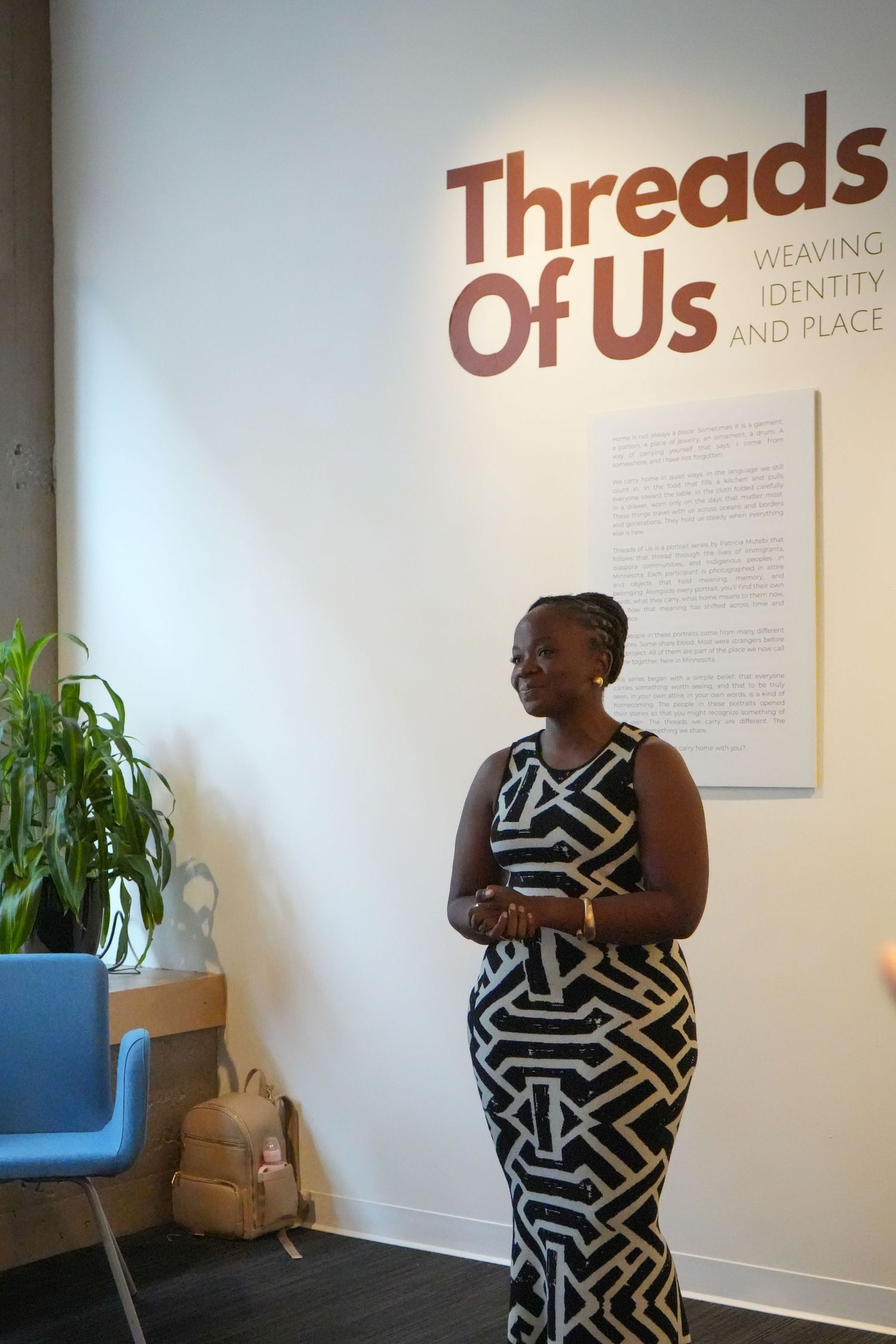 Woman in geometric dress stands beside Threads Of Us: Weaving Identity and Place exhibition sign at gallery.