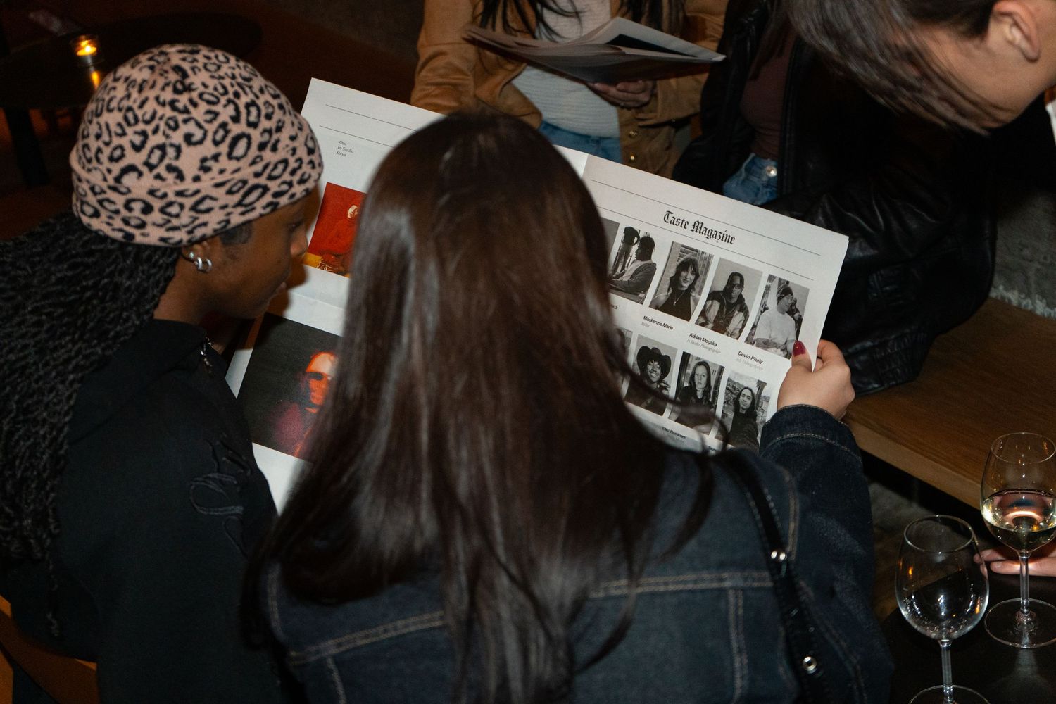 Three people looking at a magazine or program together at an indoor event, with a wine glass visible in the background.