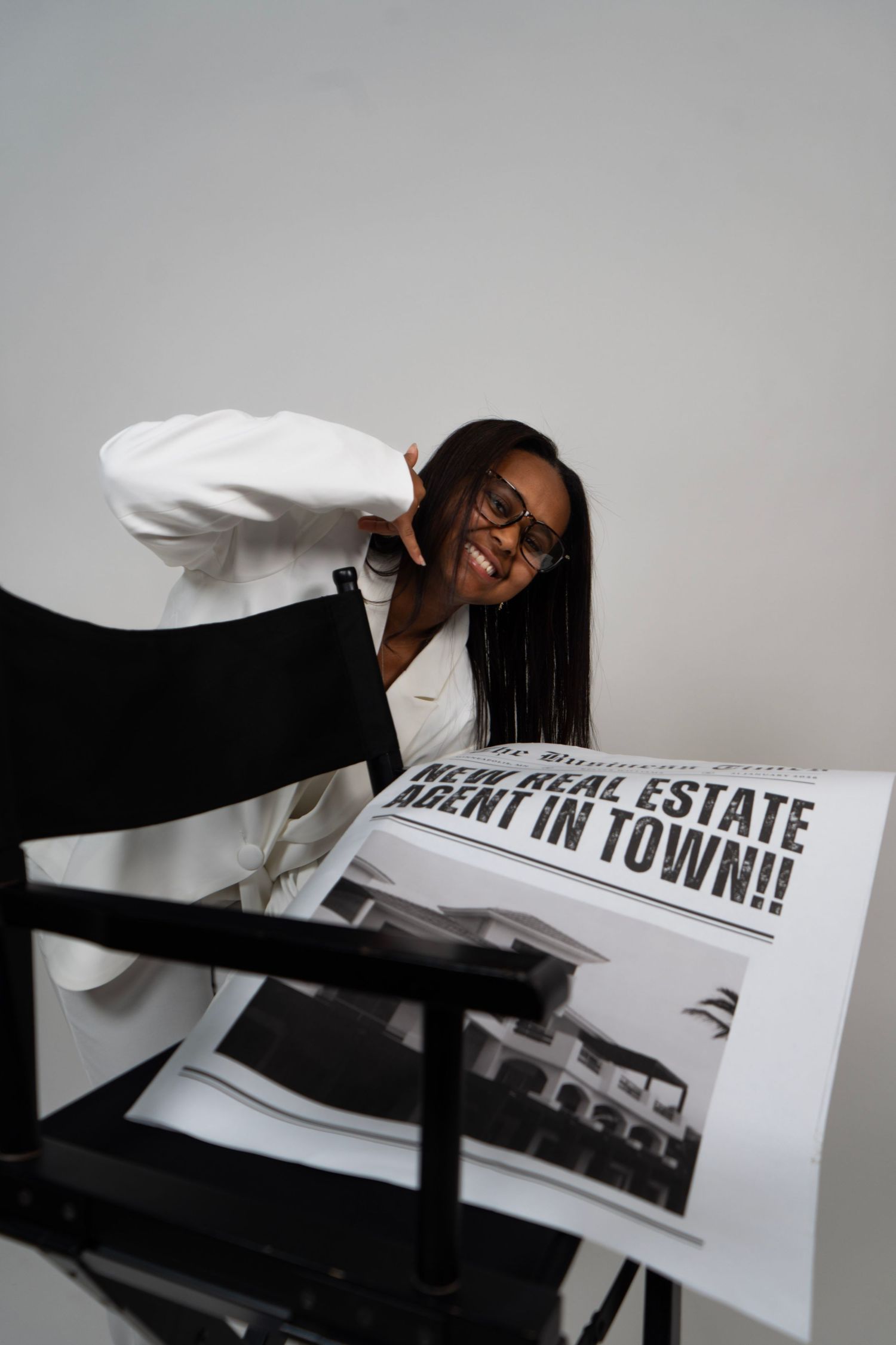 Smiling Black woman in white suit posing with a newspaper headline reading New Real Estate Agent In Town on a director's chair.