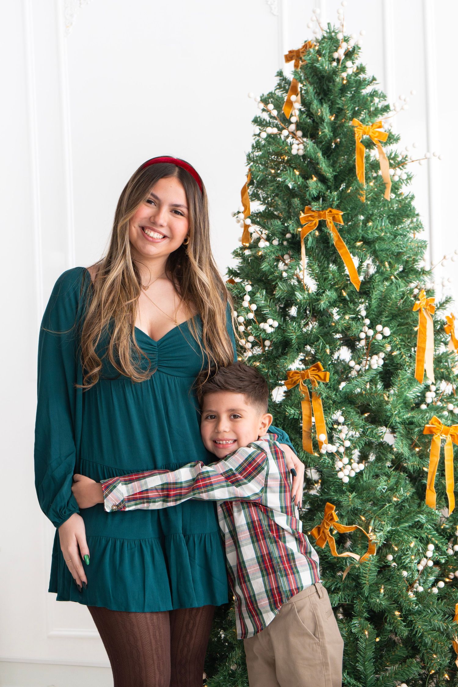A smiling woman in a teal dress hugs a young boy near a decorated Christmas tree with gold ribbon bows.