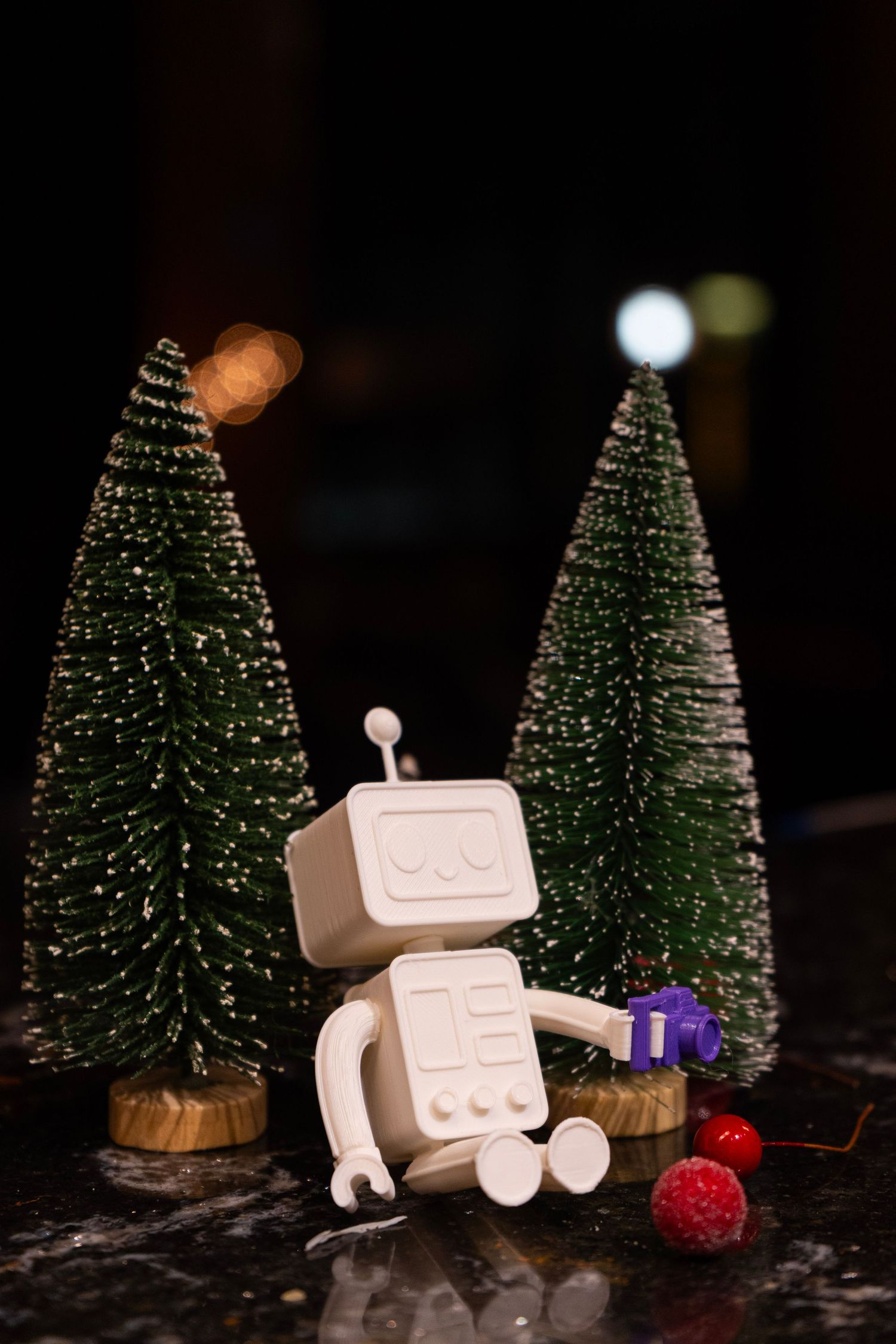 White toy robot figurine surrounded by miniature snow-dusted Christmas trees with colorful ornaments on dark surface.