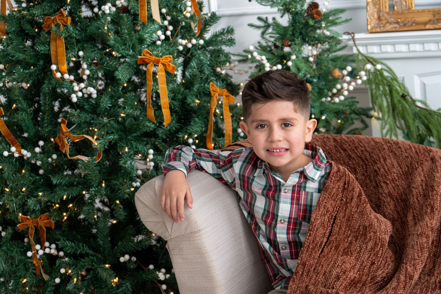 A smiling boy in a plaid shirt sits on a couch beside a decorated Christmas tree with gold bows.