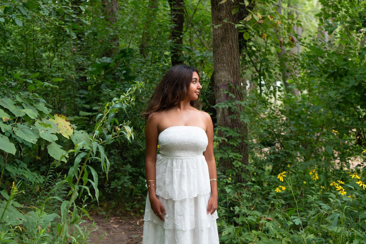 Woman in white strapless maxi dress standing on a forest path surrounded by lush green trees and foliage.