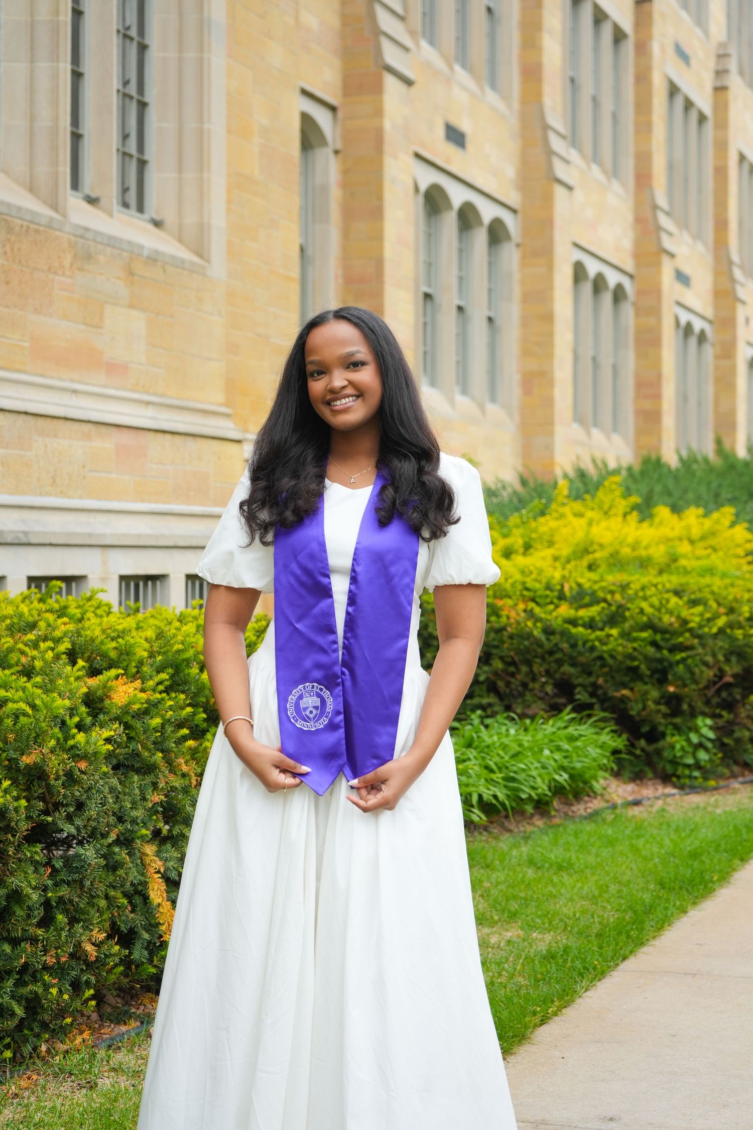 Young woman in white dress and purple graduation stole smiling outdoors near a stone building and green shrubs.