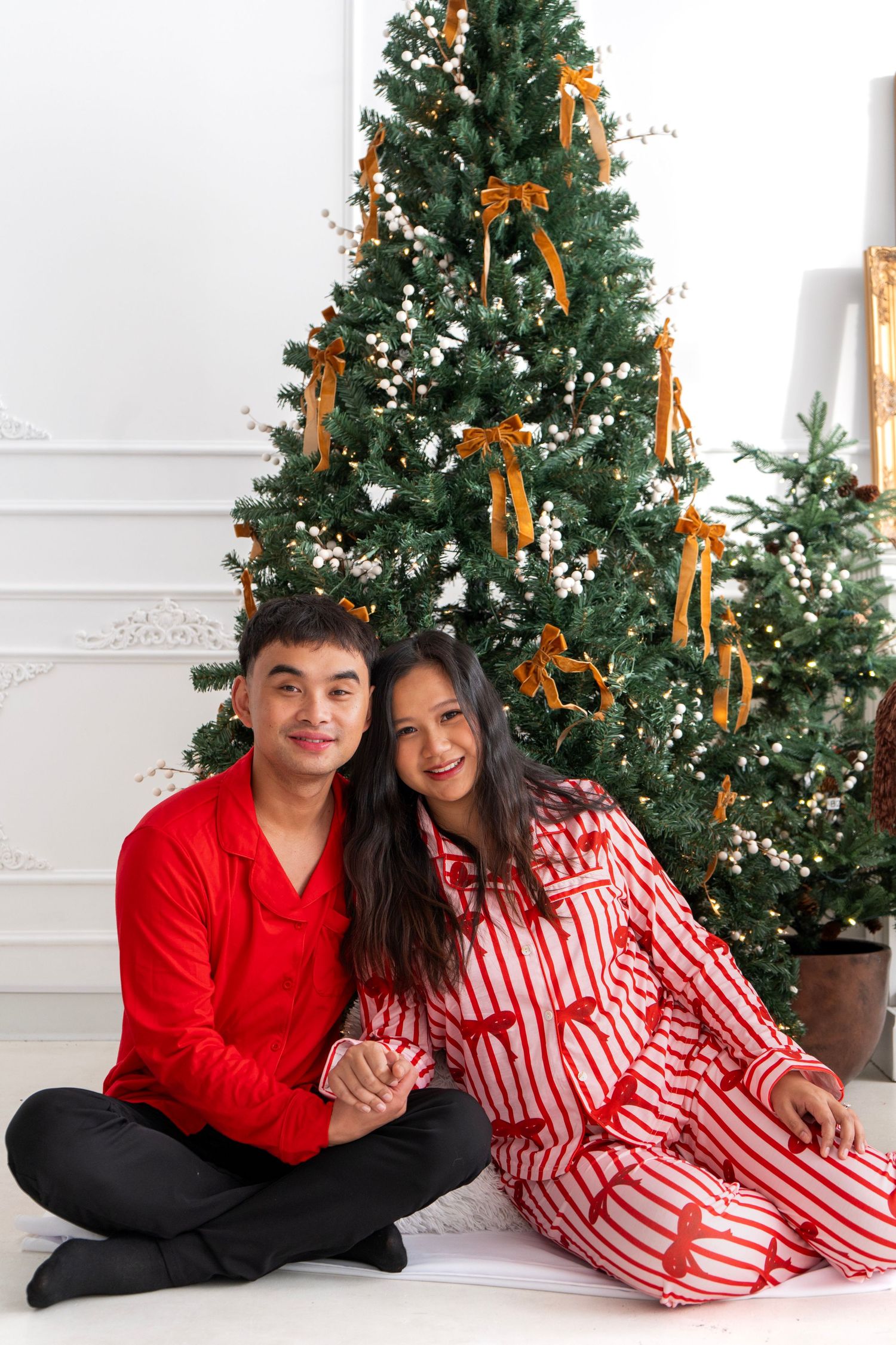 Couple in red pajamas sitting together in front of decorated Christmas tree with gold ribbons.