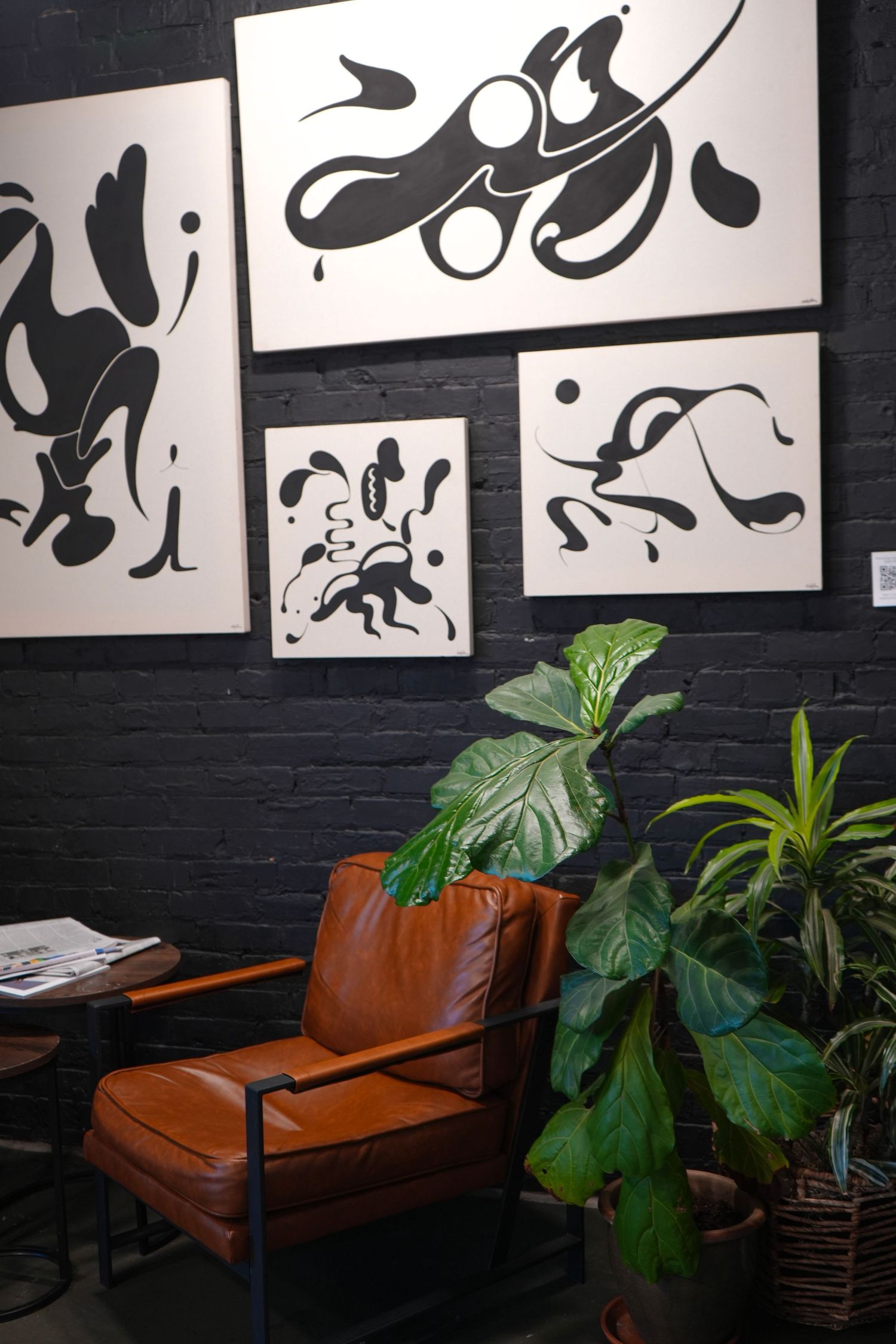 Brown leather armchair beside lush plants against dark brick wall with black and white abstract art paintings.