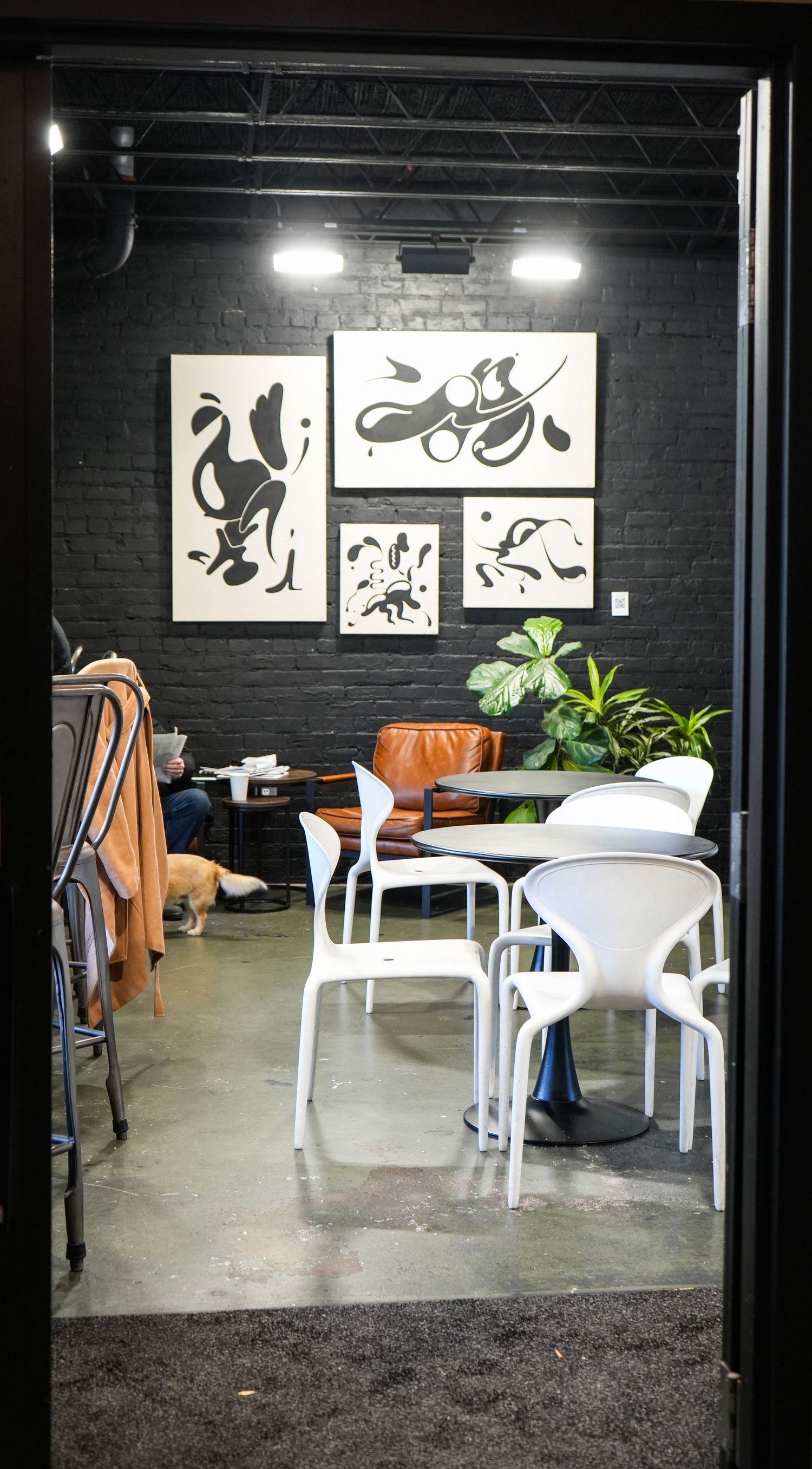 Modern cafe interior with black brick walls, white chairs, abstract art gallery wall, and a dog resting near patrons.