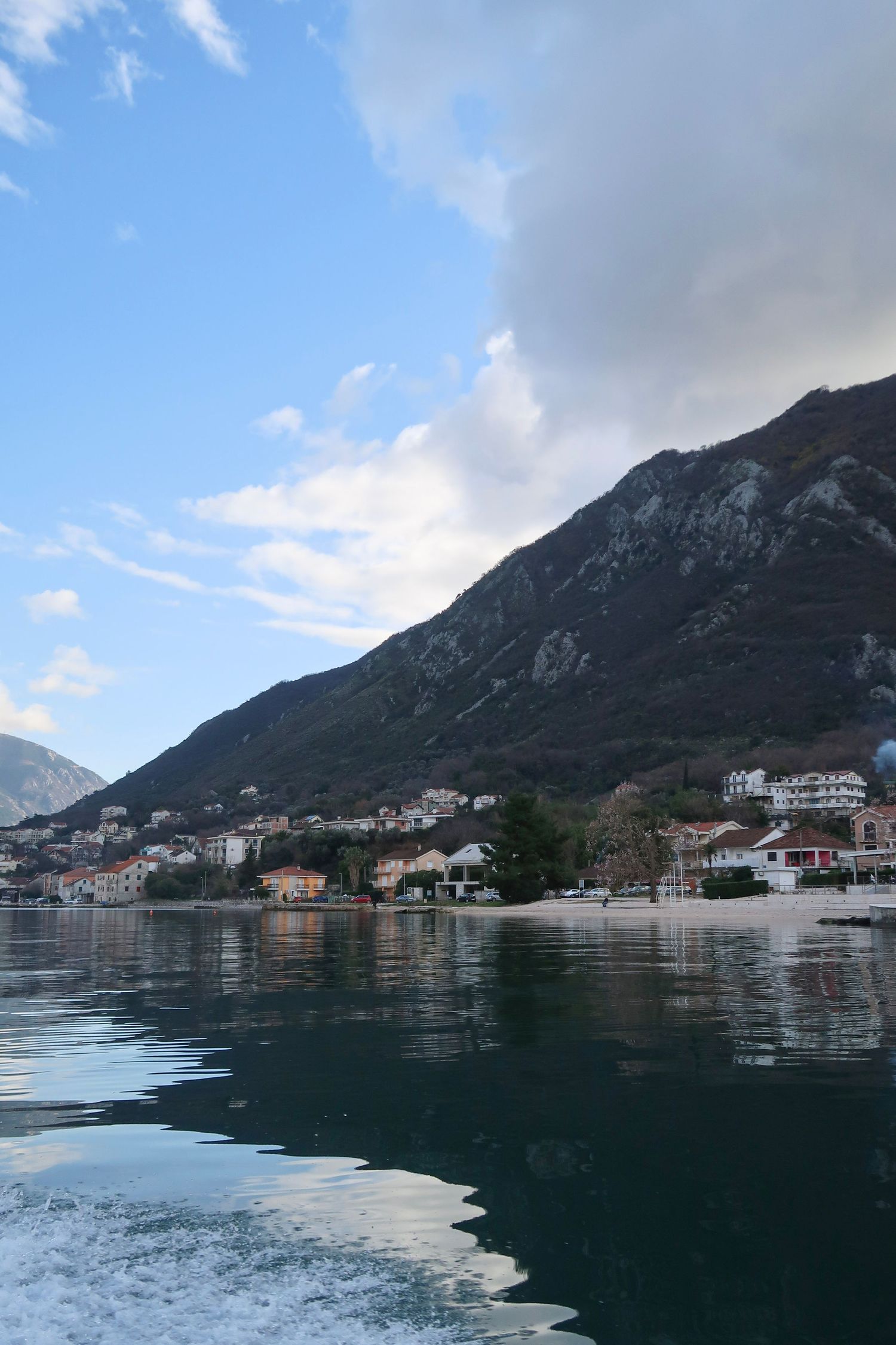 Scenic coastal town nestled at the base of a mountain, reflected in calm bay waters under a partly cloudy sky.