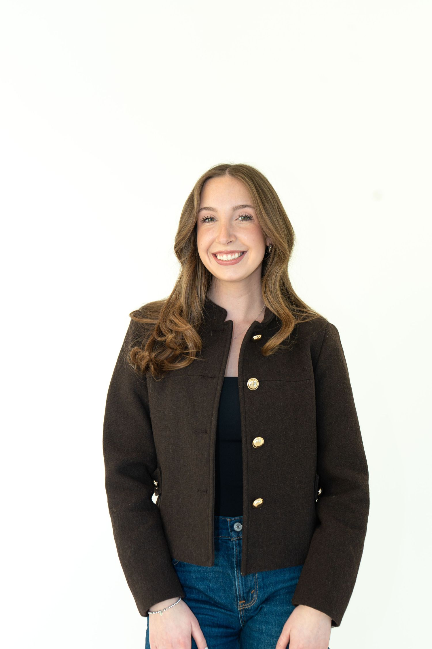 Smiling young woman in dark brown jacket and jeans posing against white background.