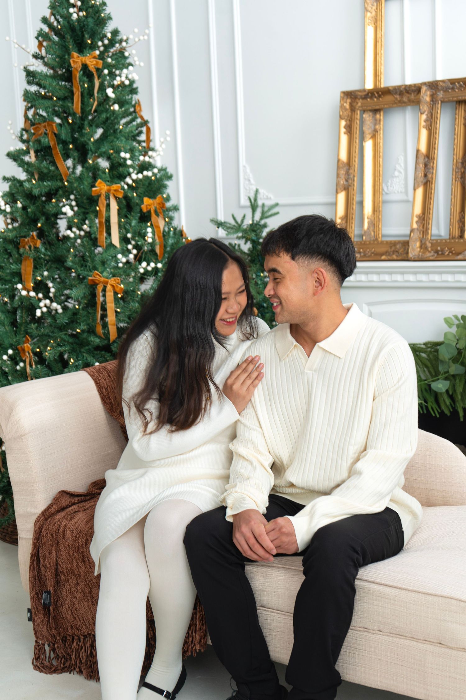 Asian couple in white sweaters sitting on sofa by decorated Christmas tree with gold ribbons.