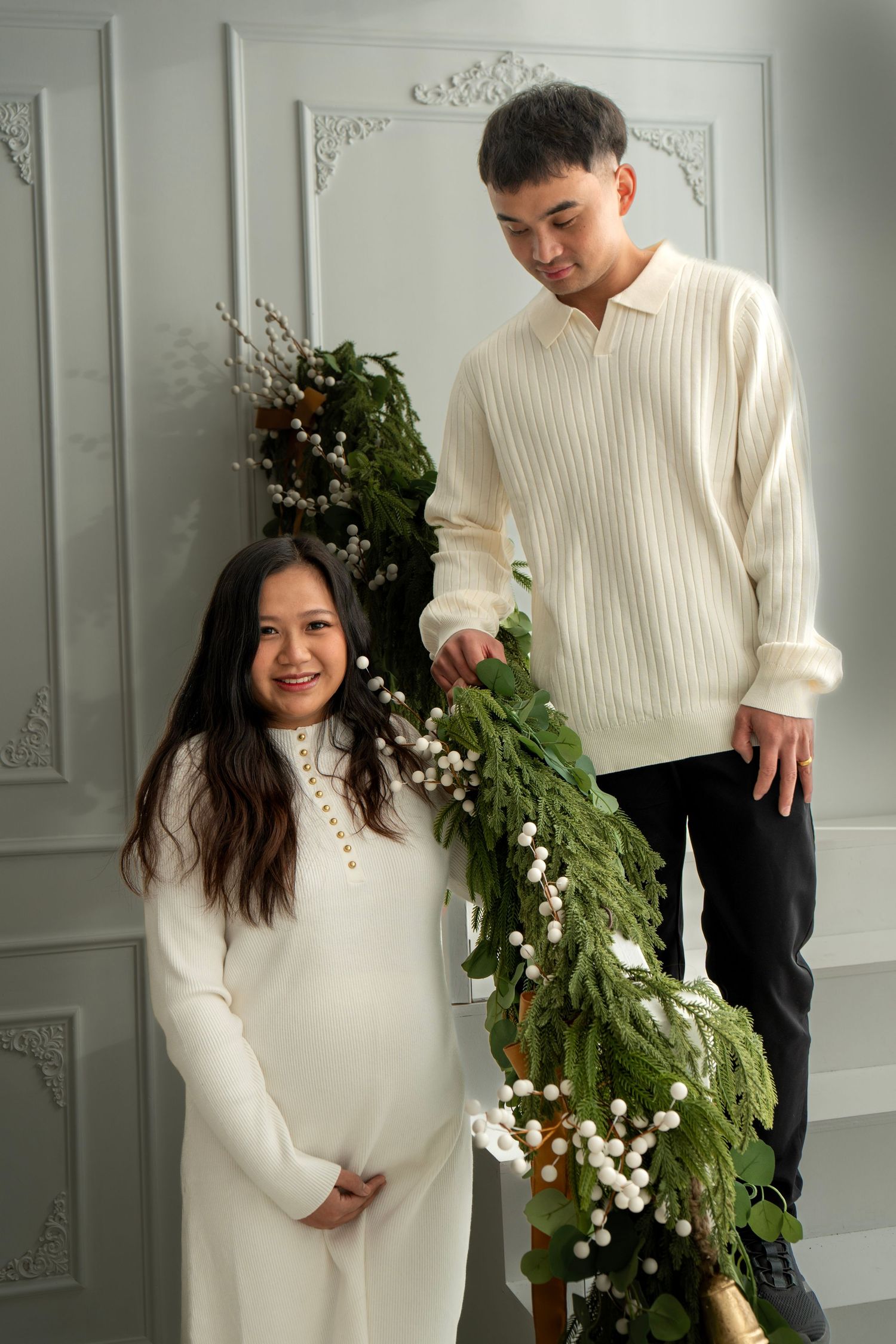 Pregnant woman and man in white outfits pose with festive green garland decor against elegant gray wall.