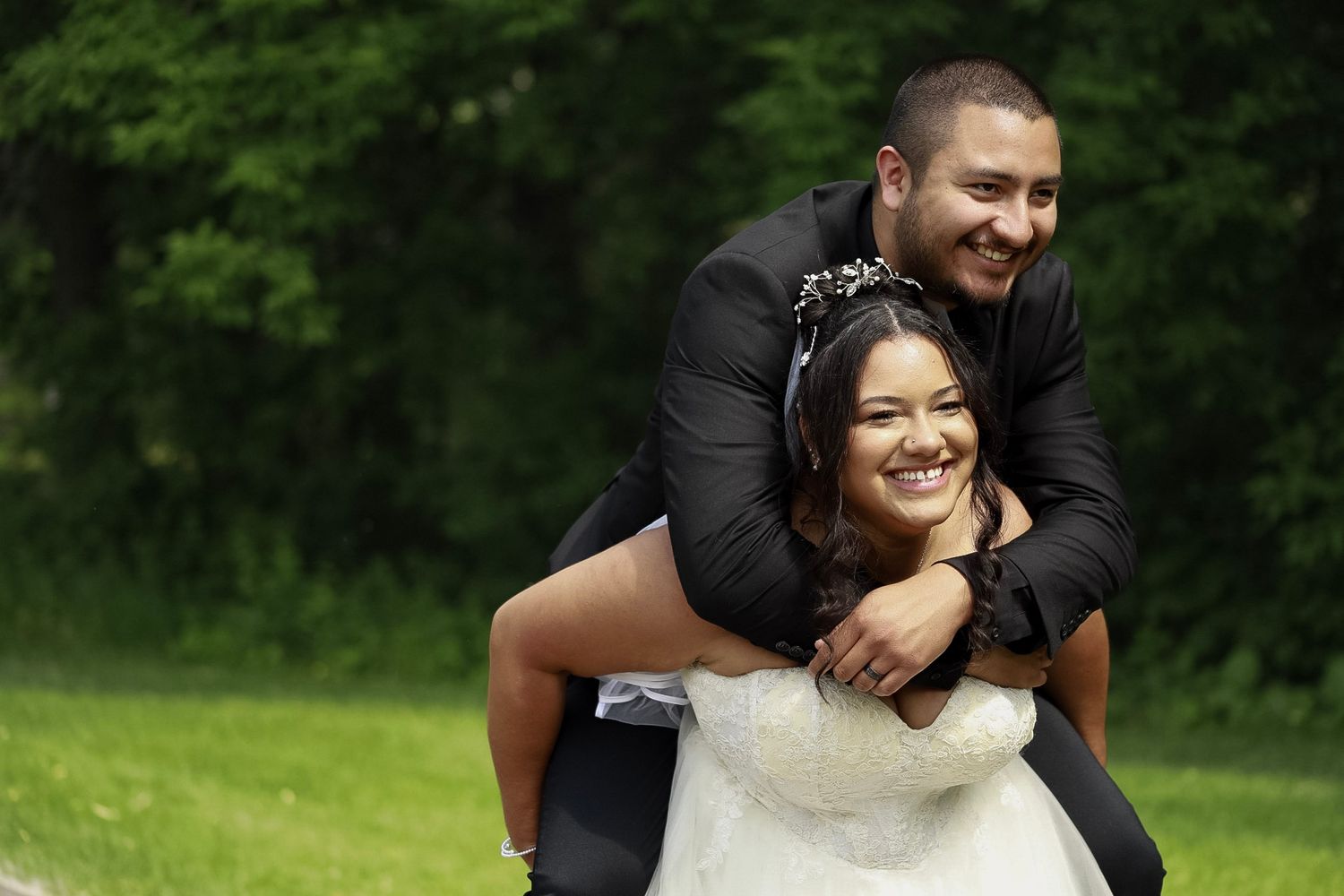 Joyful bride piggyback riding groom in black suit outdoors on green grass with trees in background.
