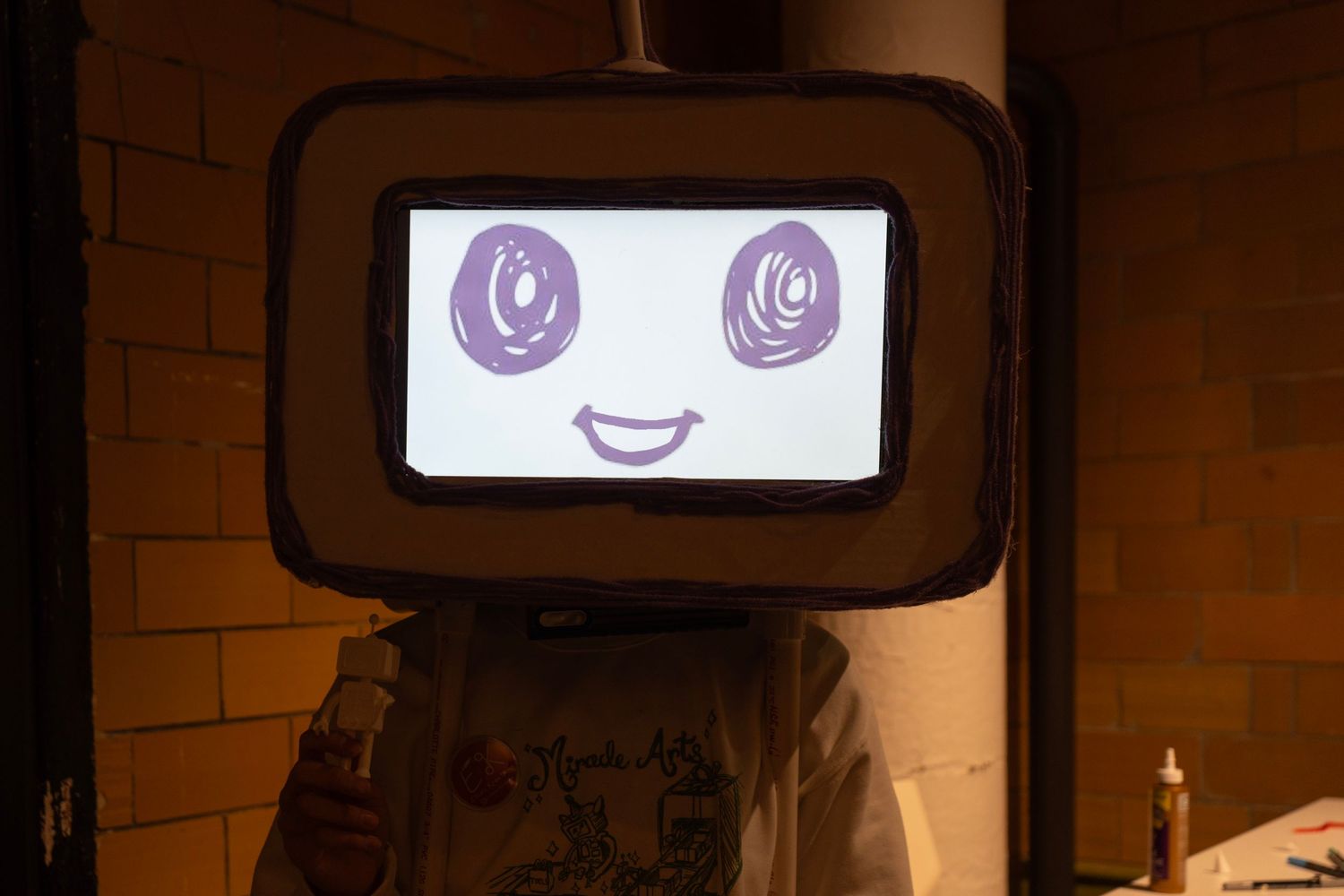 A person wearing a TV head costume with a smiling cartoon face displayed on the screen in purple ink.