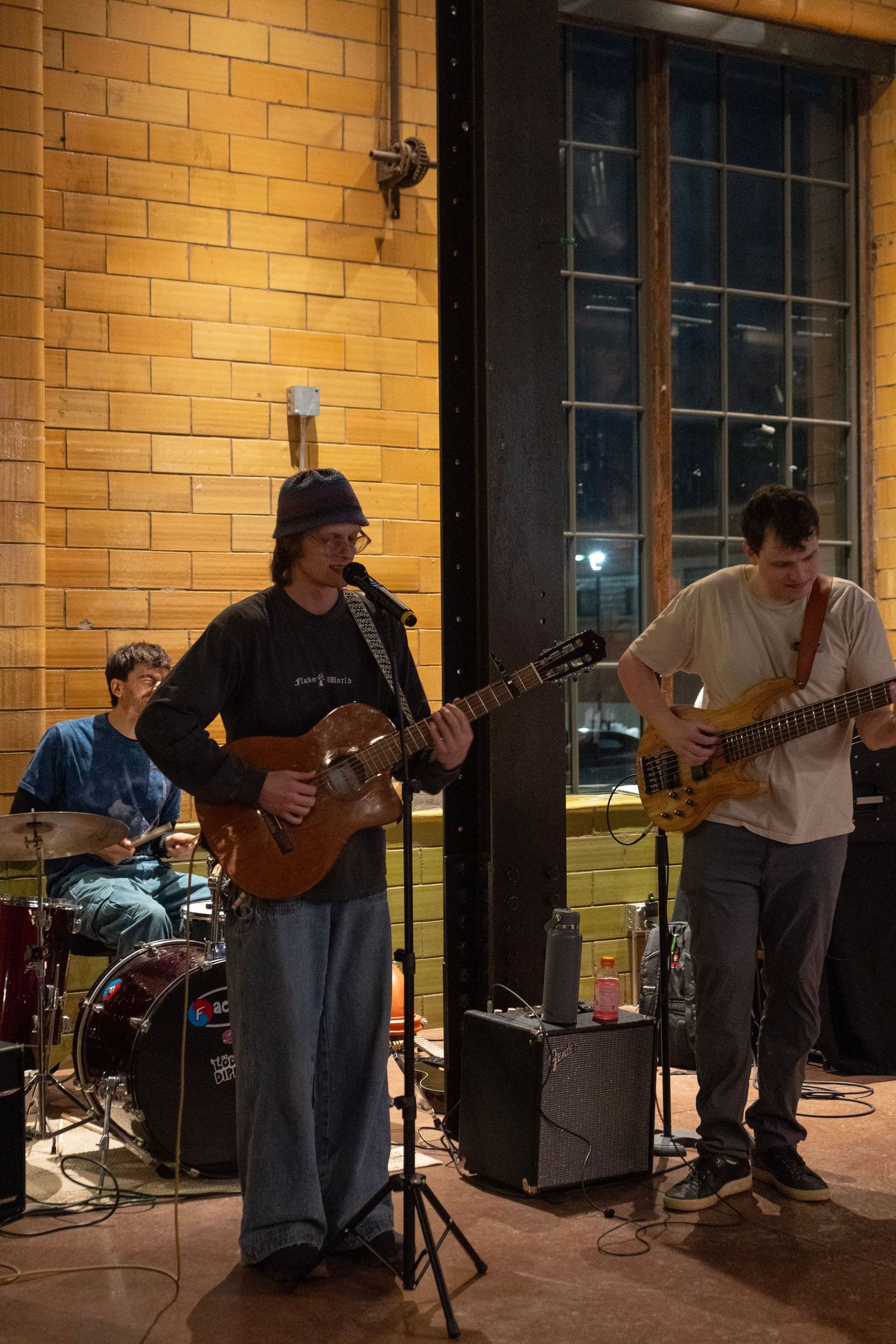 A three-piece band performs live, featuring a guitarist at mic, bassist, and drummer against a warm yellow brick wall.