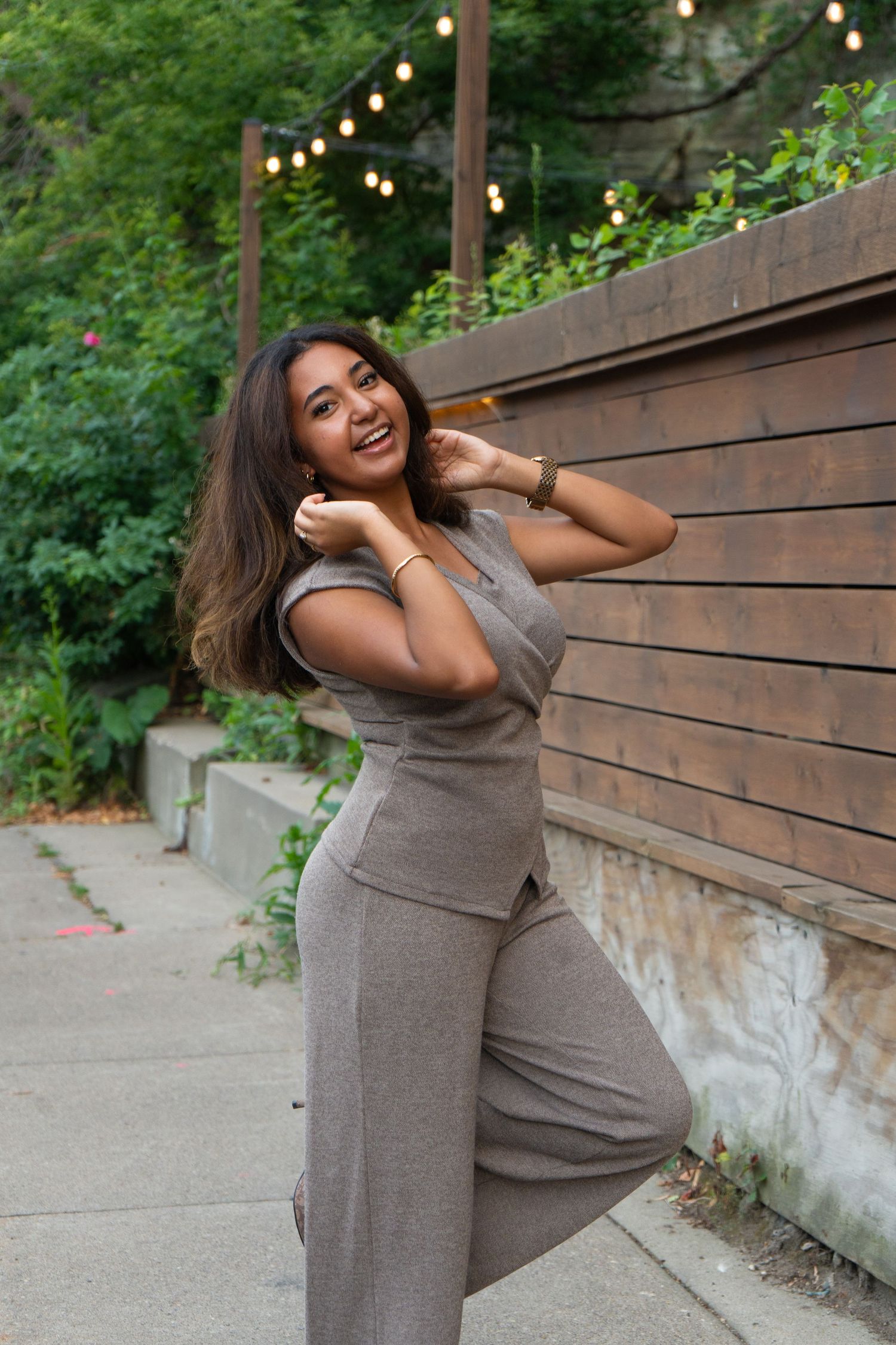Smiling young woman in gray jumpsuit posing outdoors near wooden wall with string lights and greenery.