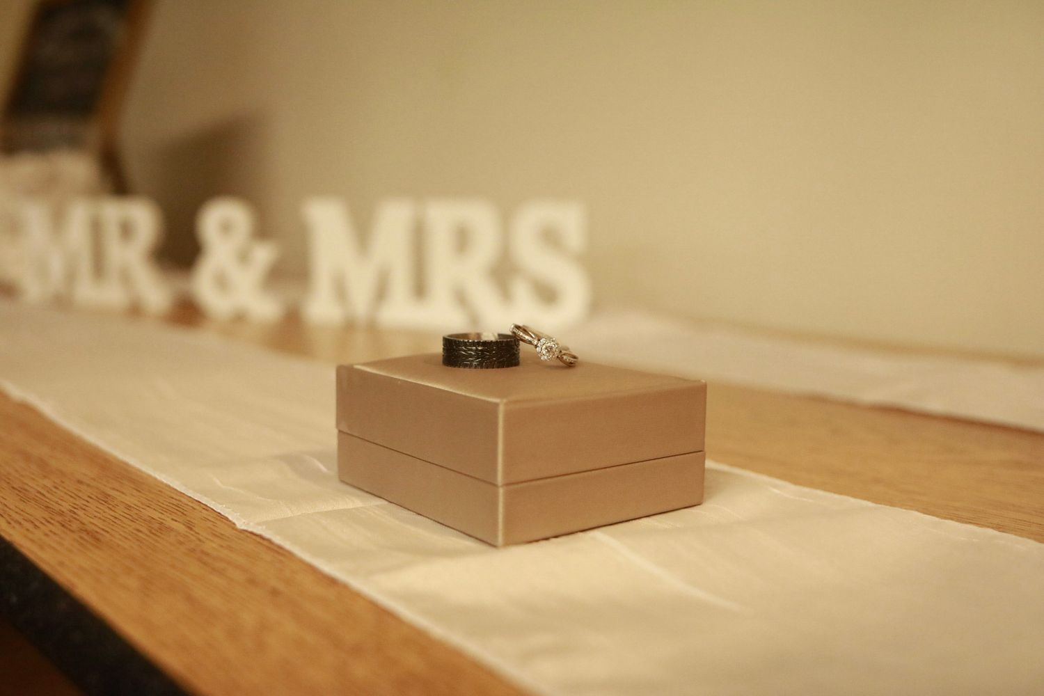 Wedding rings displayed on a beige box with MR & MRS letters in the background on a wooden shelf.