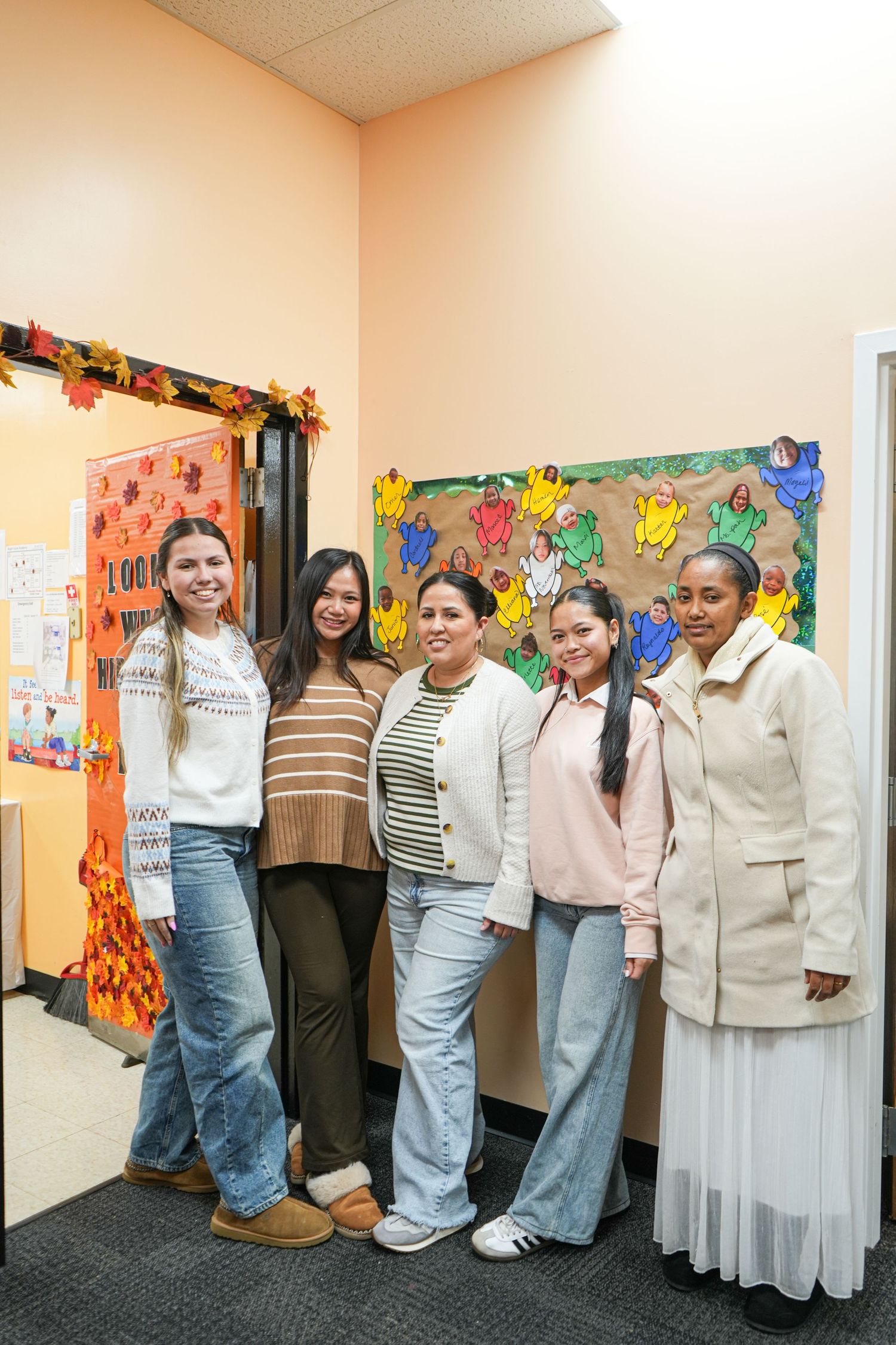 Five women smiling together in a hallway decorated with colorful fall-themed artwork and autumn leaf decorations.