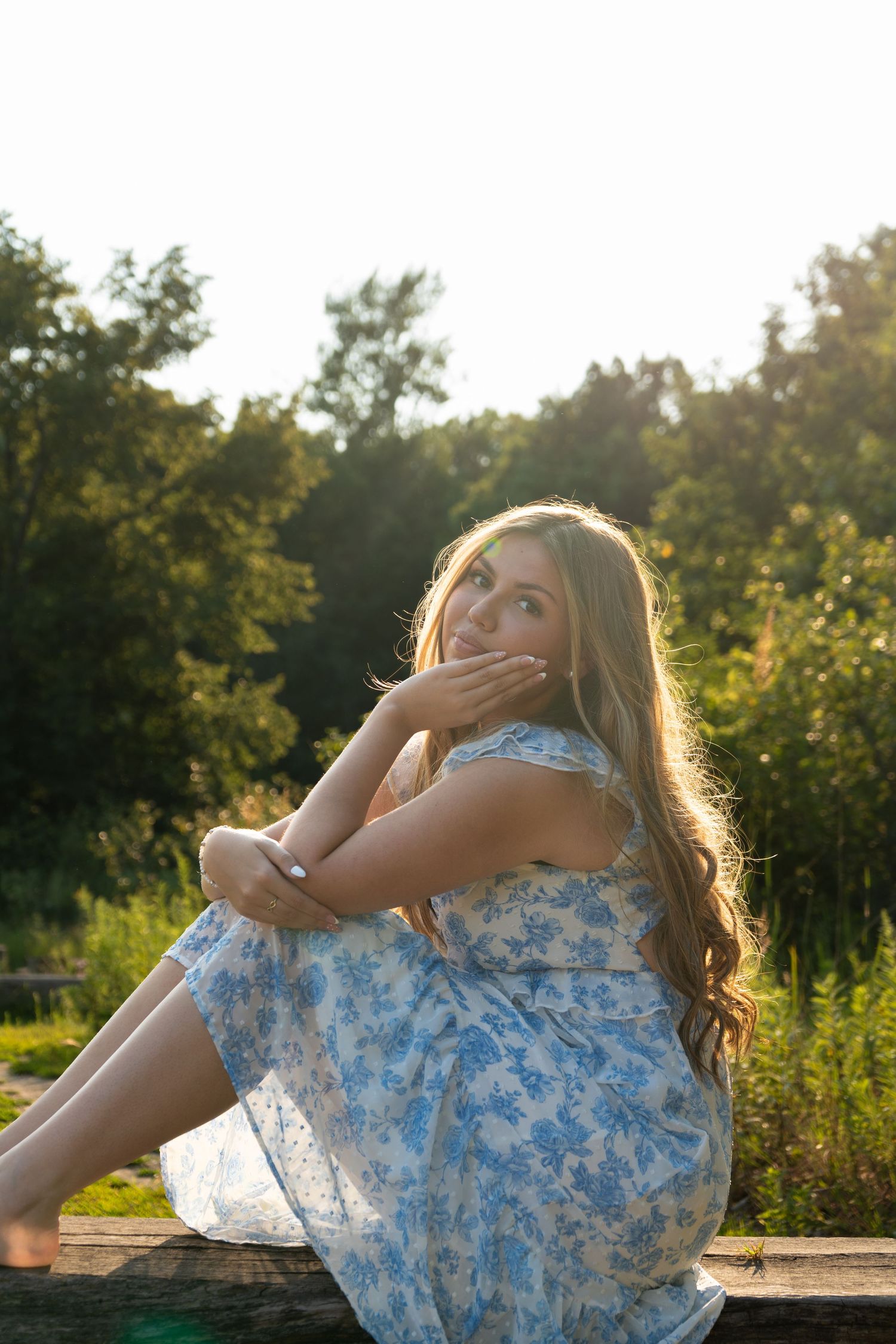 Young woman in floral blue dress sitting outdoors on wooden surface, smiling with golden sunset light filtering through green trees.