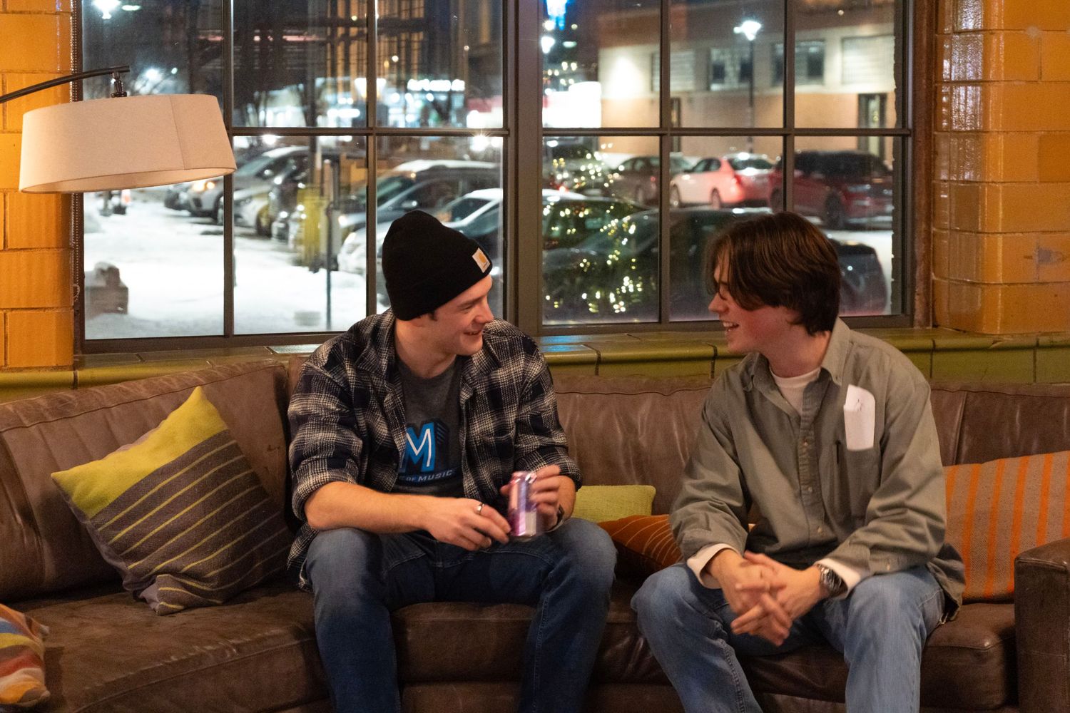Two young men sitting on a couch in a cozy cafe, smiling and chatting together at night.