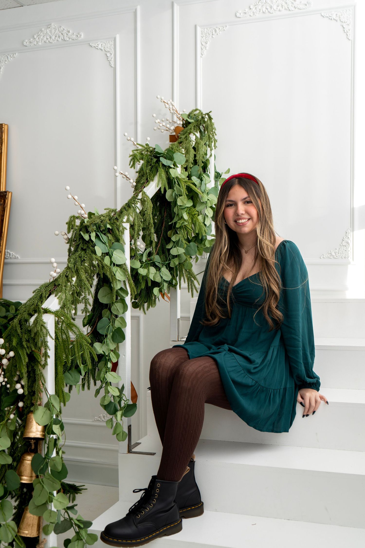 Woman in teal dress and red headband sitting on white stairs with festive green garland holiday decor.