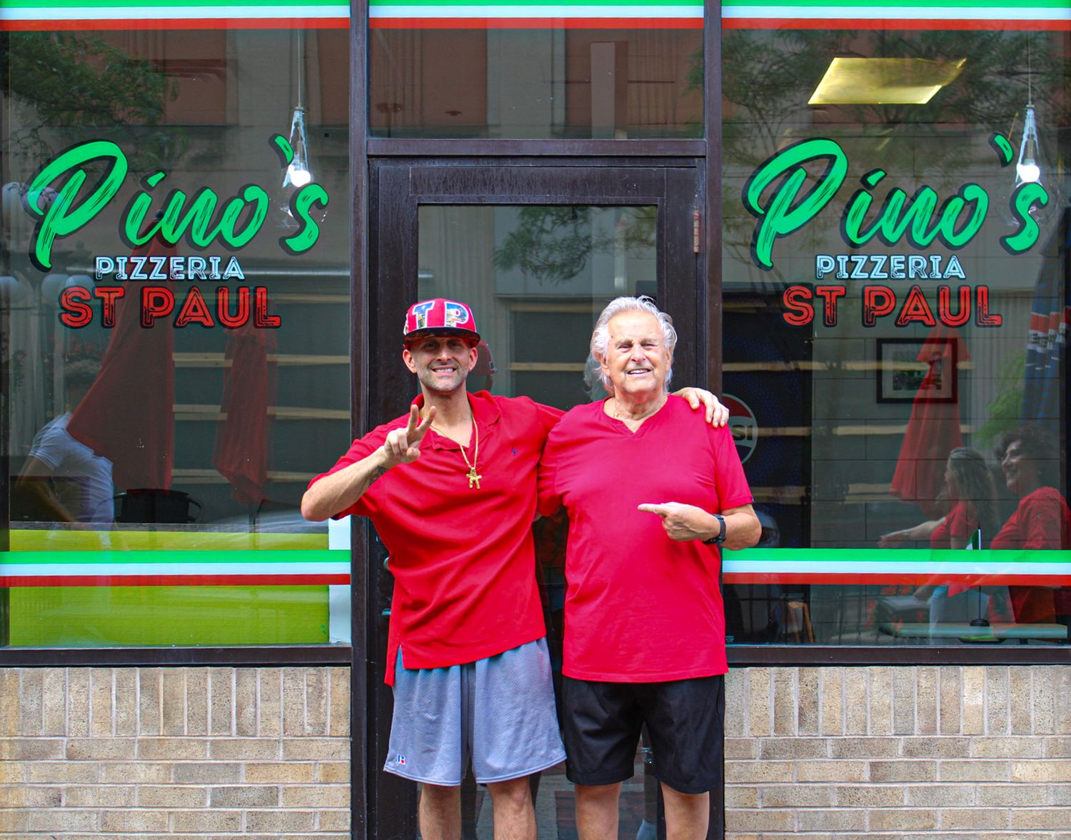 Two men in red shirts smiling in front of Pinos Pizzeria St Paul restaurant entrance on a sunny day.