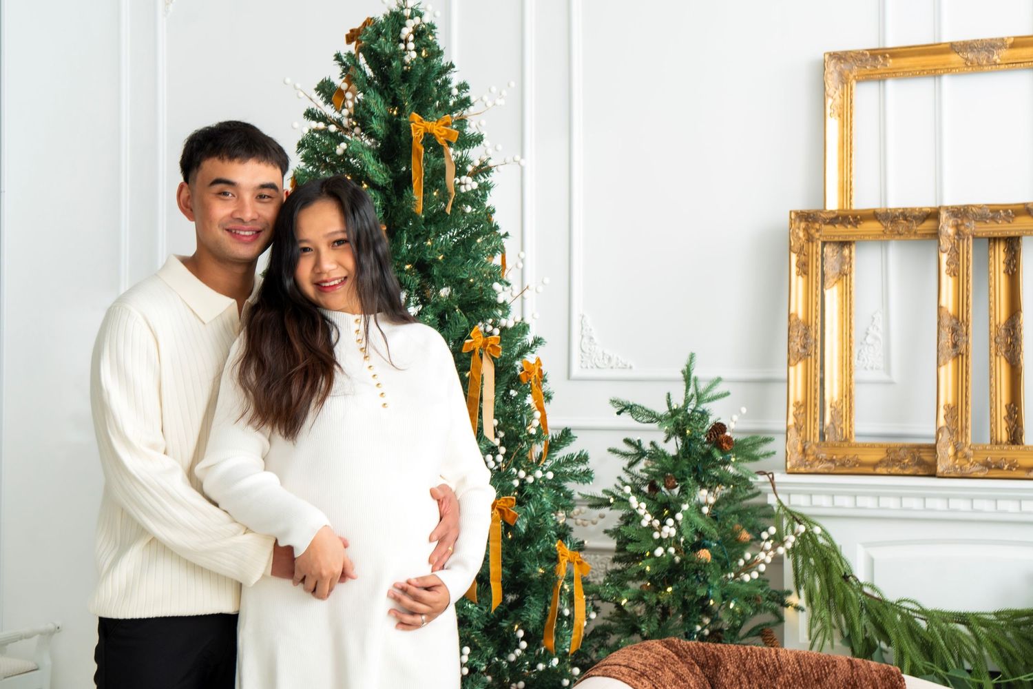 Smiling couple in white outfits embracing by decorated Christmas tree, pregnant woman cradling baby bump.