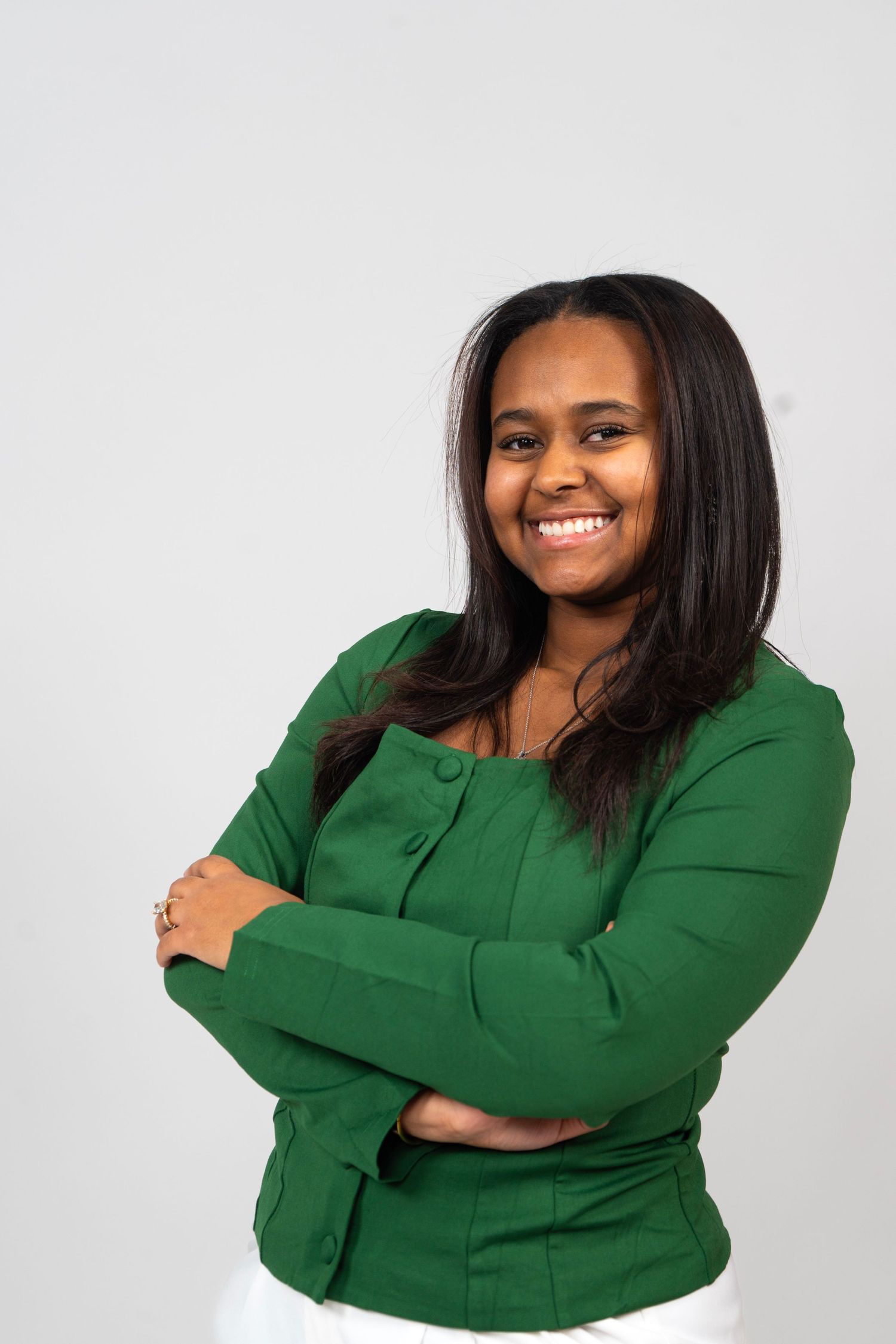 Smiling young woman in a green blazer with arms crossed against a light gray background.