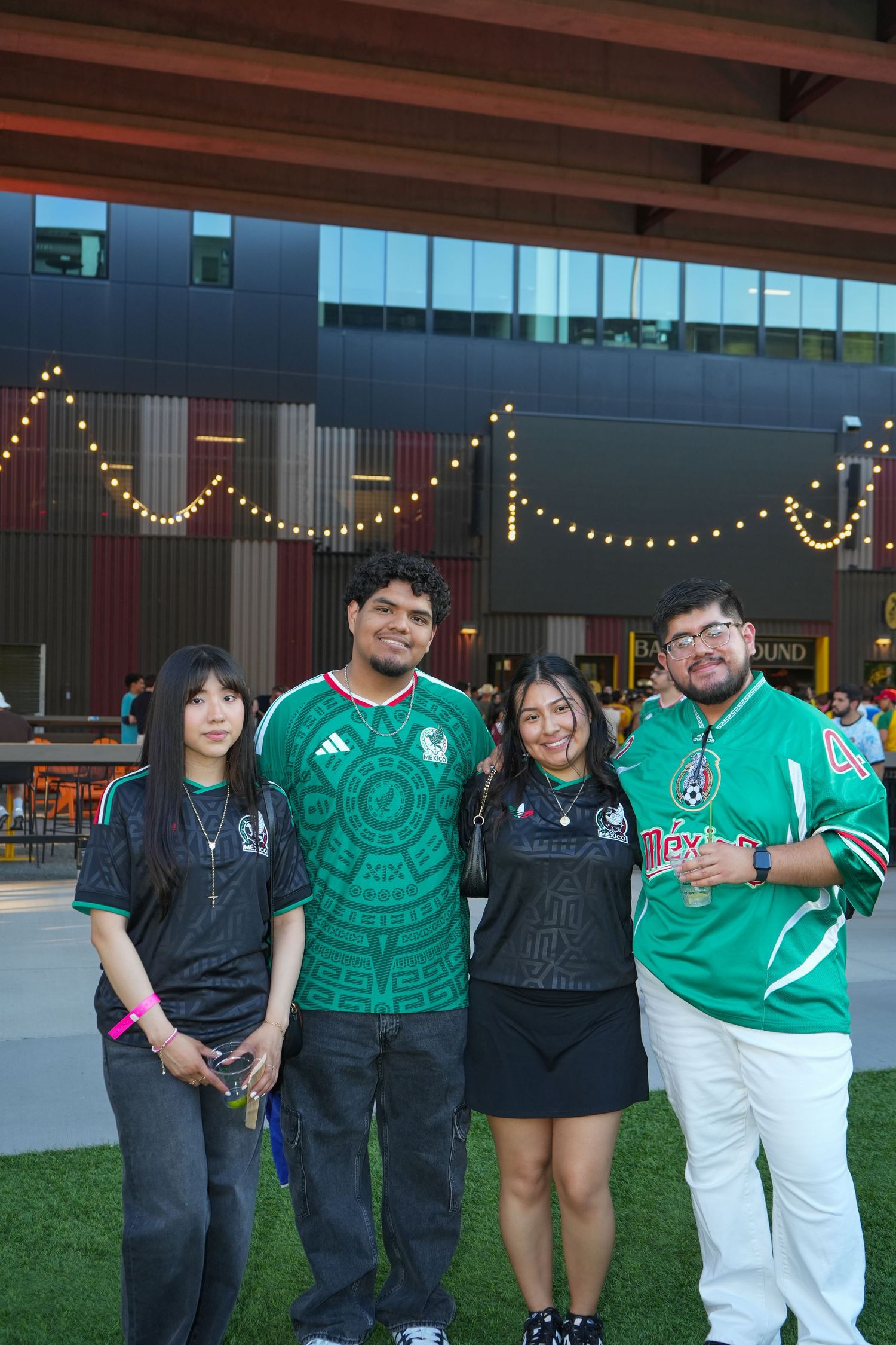 Four friends pose together outdoors wearing Mexico soccer jerseys at a festive evening event with string lights.
