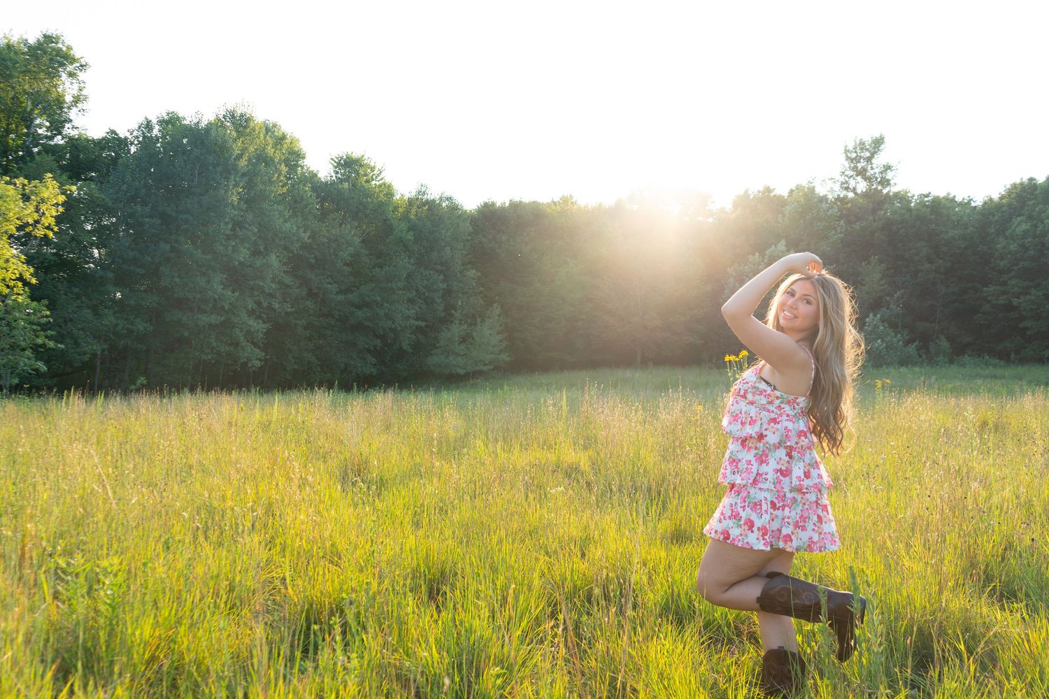 Young woman in floral dress and boots posing in sunlit green meadow with trees in background.